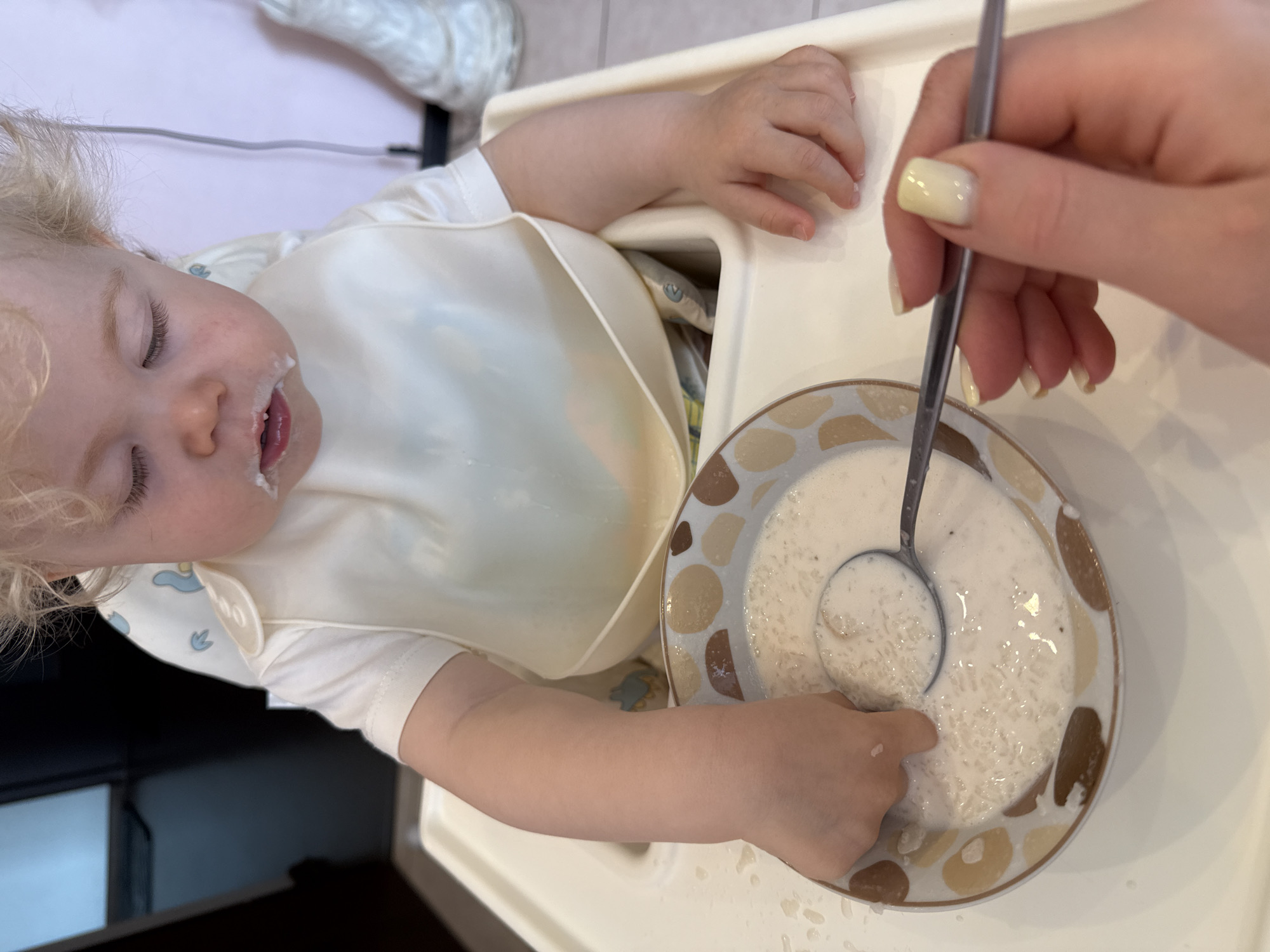 A child with light blonde curly hair is seated in a highchair, wearing a white bib and shirt. The child's eyes are closed, and there is food smeared around the mouth. The right hand is reaching into a bowl of white, creamy substance, while a spoon is held above the bowl by an adult hand. The adult's fingernails are painted a pale yellow color. The bowl has a pattern of beige and brown circles around the rim. The scene appears to be indoors, with a beige highchair and a kitchen or dining area in the background. The exact location is Unknown, Unknown.