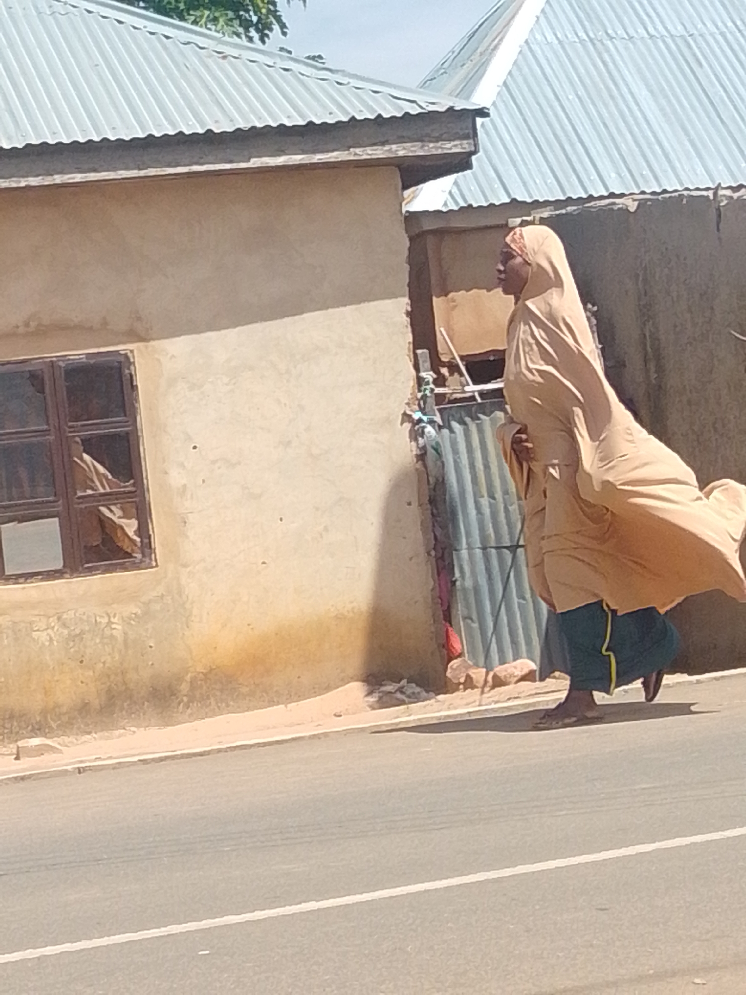 A person wearing a tan headscarf and a dark green skirt walks on the right side of a paved road in an outdoor, likely rural, environment. The person is walking away from the camera, and their face is not clearly visible. Behind the person, there is a tan-colored building with a corrugated metal roof. A window with brown trim is visible on the side of the building. To the right of the building, there is another corrugated metal structure. The road has a white line marking the lane. The sun is shining, casting shadows on the ground.