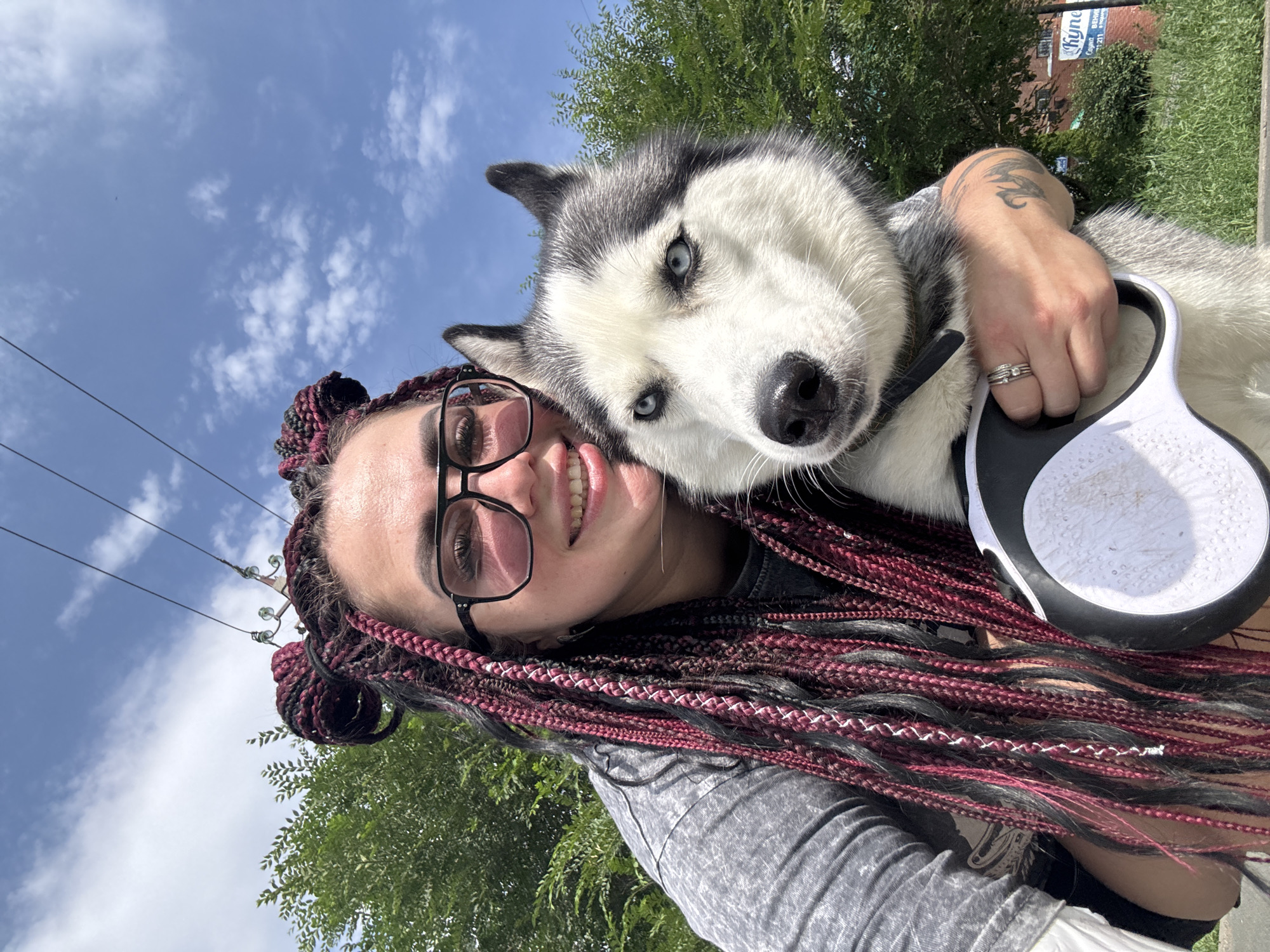 In Tolyatti, Russia, a woman with long, dark and burgundy braided hair and large-framed glasses smiles as she takes a selfie with her husky. The husky, with striking blue eyes and black and white fur, is held close by the woman. She is wearing a grey t-shirt and a ring on her left hand. The dog wears a collar and a retractable leash handle is visible. The background features a bright blue sky with scattered clouds, overhead power lines, lush green trees, and a glimpse of buildings with signage, including one with Russian text.