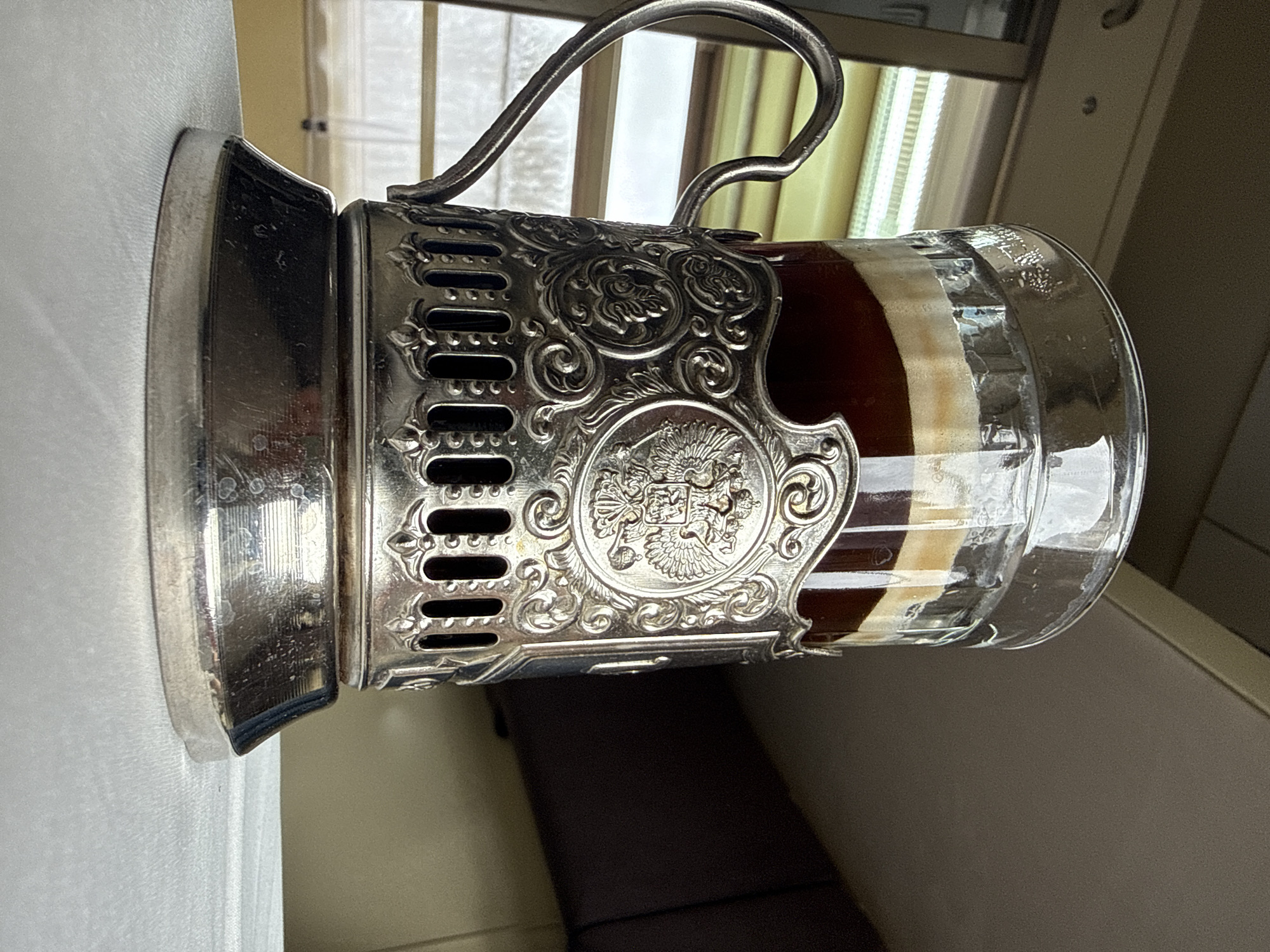 A glass of layered liquid, appearing to be coffee or a similar beverage, is held in a silver-colored metal podstakannik (cup holder). The podstakannik features decorative filigree with a prominent emblem in its center. The handle of the podstakannik curves upward and back. The scene appears to be indoors, near a window covered by thin, light-colored curtains. Part of a seat or bench with a brown cushion is visible below the cup. The location is purportedly Vyglyadovka, Russia.