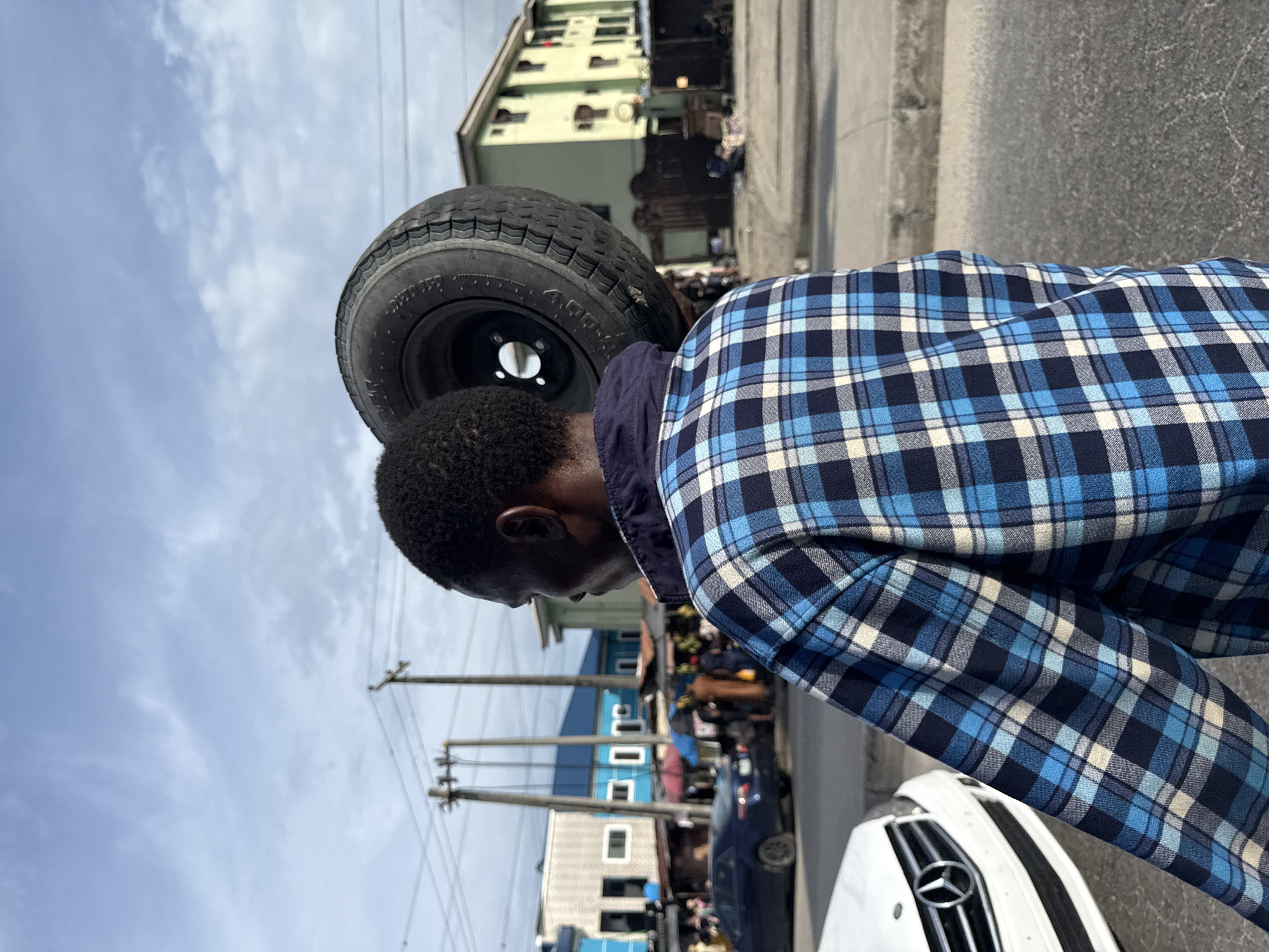 A side-view, medium shot shows a person with dark skin wearing a blue plaid shirt, carrying a tire on their shoulder. The location is Ado, Nigeria, as evidenced by the urban environment, the type of buildings, the power lines, the presence of a Mercedes-Benz car and the other individuals in the background, suggesting a marketplace setting. The image is oriented so that the sky and buildings are visible in the background.