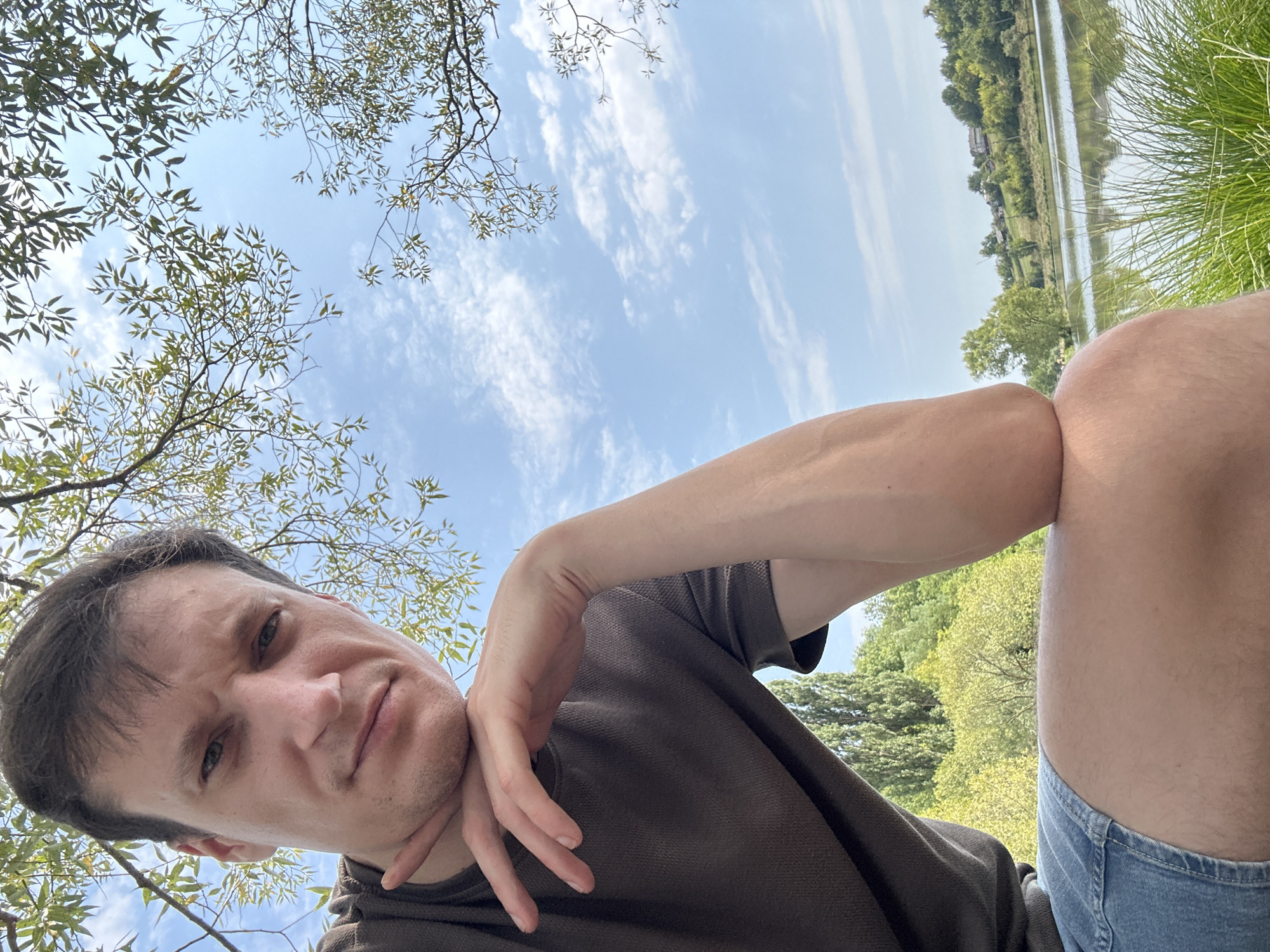 A young man with short brown hair and light skin is captured in a close-up selfie outdoors, positioned in the foreground with his chin resting on his left hand. He is wearing a dark brown polo shirt and blue denim shorts. His left arm is bent at the elbow, supporting his head. The background reveals a serene natural landscape with a calm body of water reflecting the sky. Lush green trees and vegetation flank the water, and in the distance, a gentle slope is dotted with houses, suggesting a rural or semi-rural setting. The sky above is a bright blue with scattered white clouds, partially obscured by the branches and leaves of a tree in the upper portion of the frame. The overall lighting indicates it is daytime, likely a sunny or partly cloudy day. The image appears to have been taken in the vicinity of Makiv, Ukraine, given the typical Ukrainian landscape.