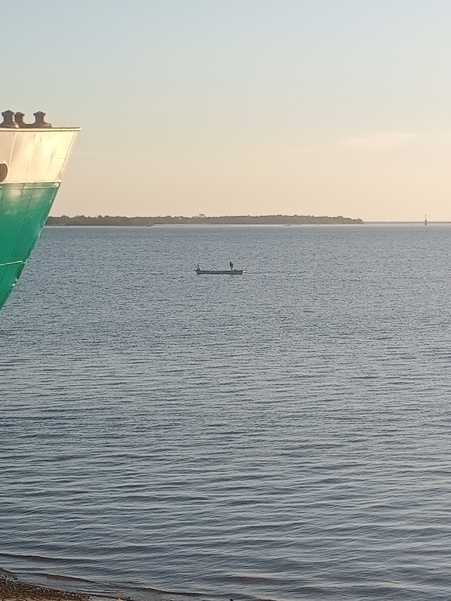 A long shot captures a calm body of water under a clear, pale sky. In the foreground, the green hull and white superstructure of a large ship occupy the left side of the frame. The water is a deep blue-gray, with gentle ripples creating horizontal lines. In the middle distance, a small, dark boat with two figures stands out against the water. The figures appear to be standing or fishing. A distant, tree-lined island stretches horizontally across the horizon, its darker silhouette contrasting with the bright sky. A small, white navigational buoy is visible to the right of the island. The image's perspective suggests it was taken from a slightly elevated position, perhaps from another boat or a pier, looking out towards the open water and the distant land.
