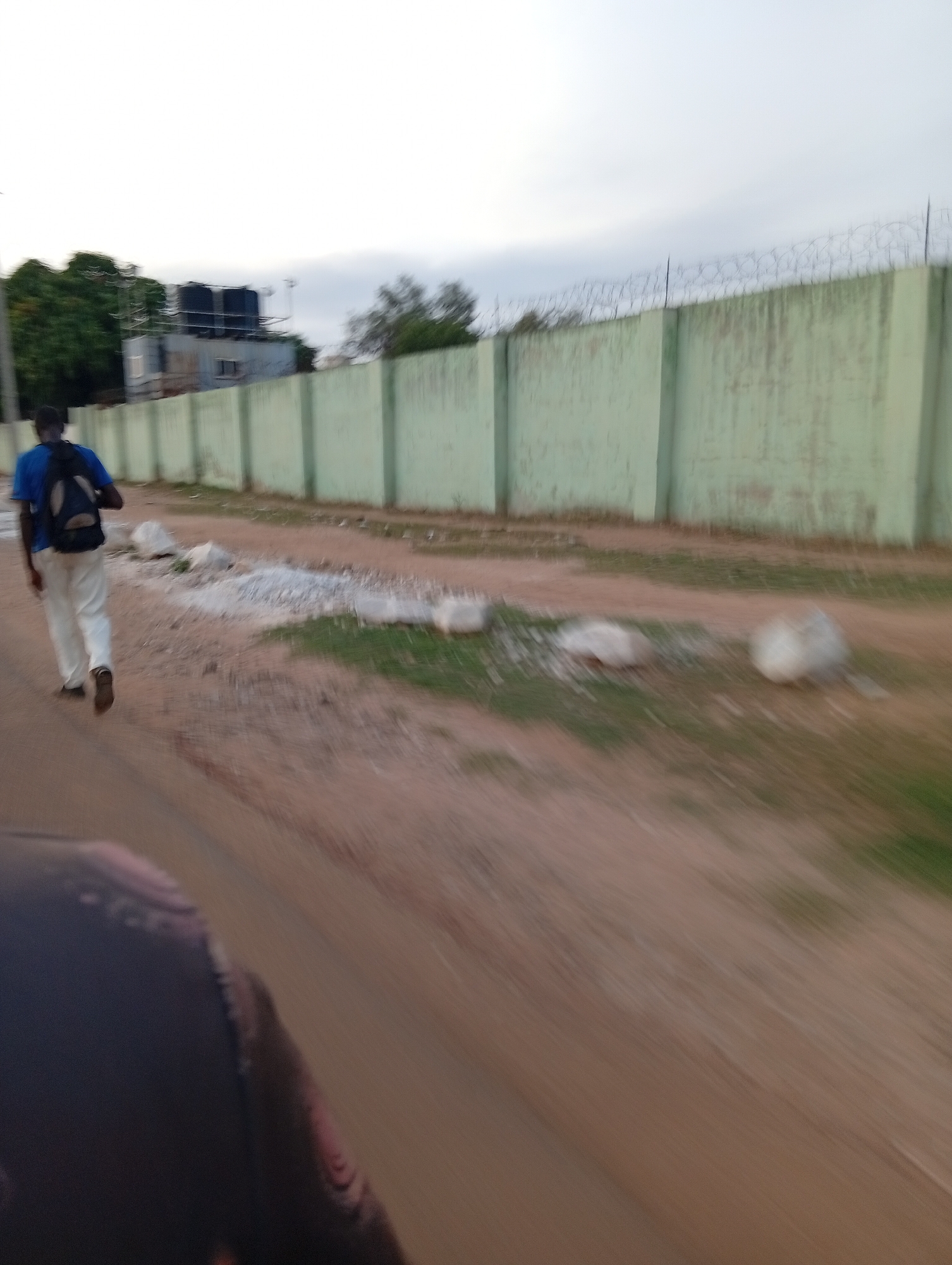 A man wearing a blue shirt, beige pants, and a dark backpack walks on a dirt road. A long, light green wall with barbed wire on top borders the road. Patches of grass and scattered rocks are visible along the side of the road. In the background, there's a building with dark water tanks on the roof, surrounded by trees. The image seems to be taken in Nassarawa, Nigeria, based on the provided information. The scene appears to be on the outskirts of a residential or industrial area. A portion of what is likely the interior of a car is visible in the bottom of the image.