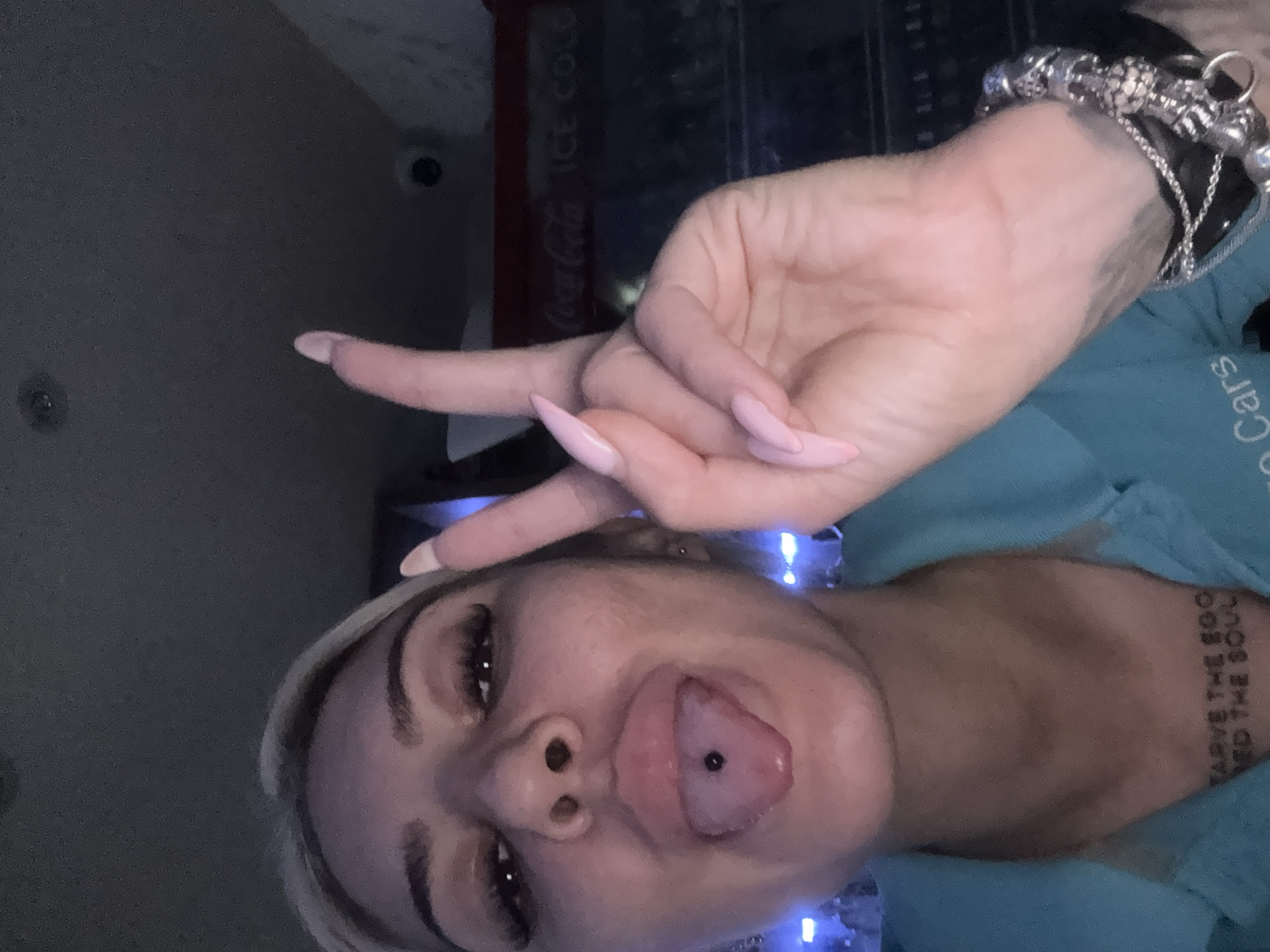 A woman is taking a selfie. She is making a peace sign with her hand. She has long, light pink fingernails and a bracelet on her wrist. The woman has blonde hair and a pierced tongue. A Coca-Cola refrigerator is in the background. The location is unknown. The tattoo on her chest cannot be determined.