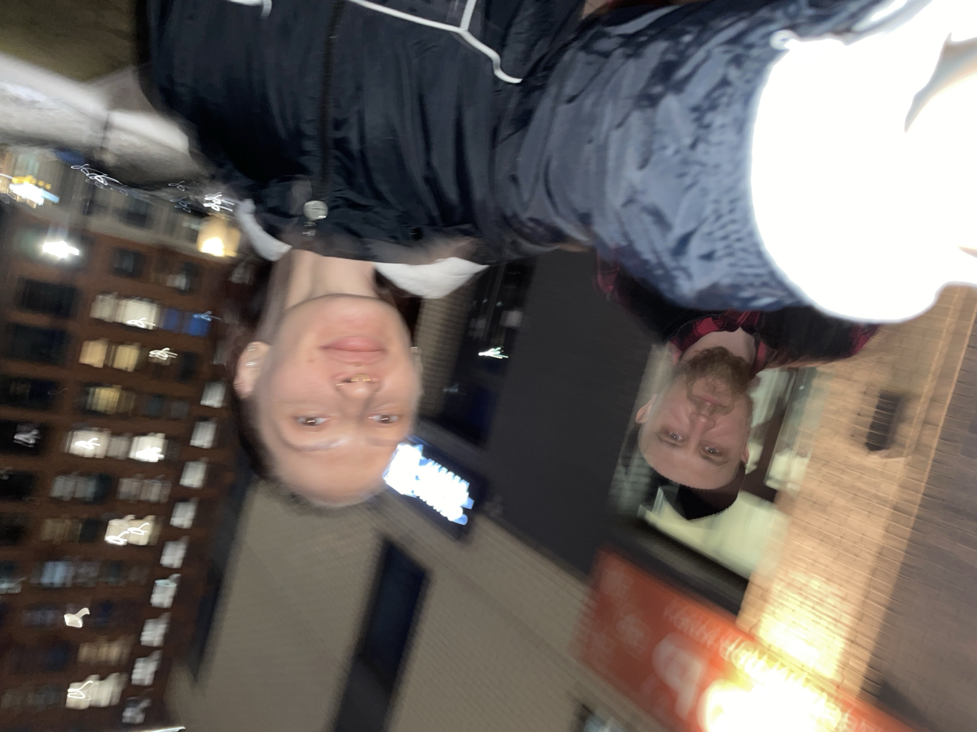 Two people are captured in the image. One person is wearing a black jacket with white trim, and the other is wearing a red and black plaid shirt with a black hat. They appear to be outside at night in a city environment.
A building with several windows illuminated can be seen in the background. A sign with bright lights and a brick building are also present.
The photo is likely taken in Izhevsk, Russia, given the location request. However, specific architectural details or signage would be necessary for a definitive confirmation.