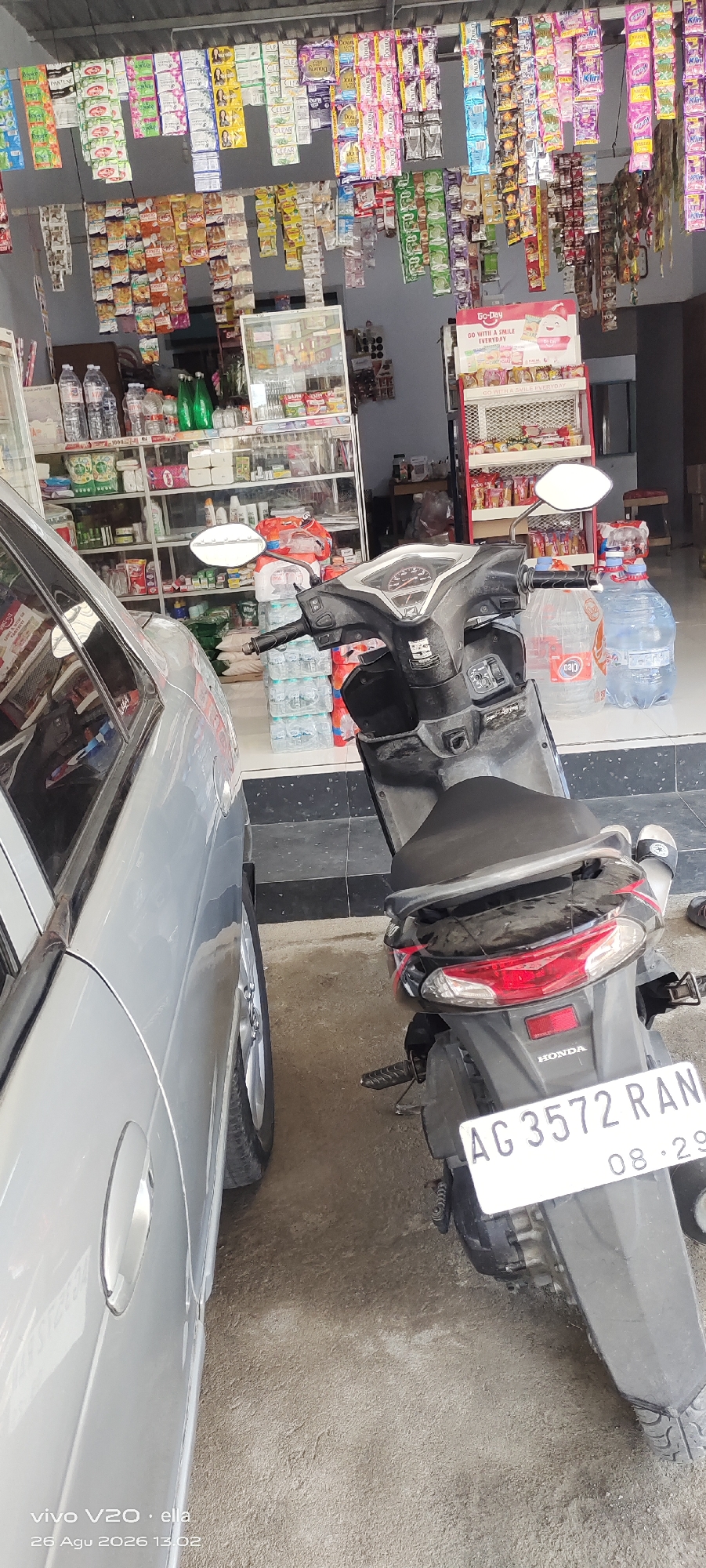This image depicts the exterior of a small shop, likely a minimarket or warung, in Simo, Indonesia. The shop is stocked with a wide variety of packaged goods, including food items, beverages, and household products, displayed on shelves and hanging from the ceiling. Two vehicles are partially visible: a silver car on the left and a black scooter on the right. The scooter is parked in front of the shop, and its license plate, "AG 3572 RAN," with a date "08.29," is clearly legible. The ground surface appears to be a mix of concrete and dirt. The overall scene suggests a typical commercial setting in a local Indonesian community.
