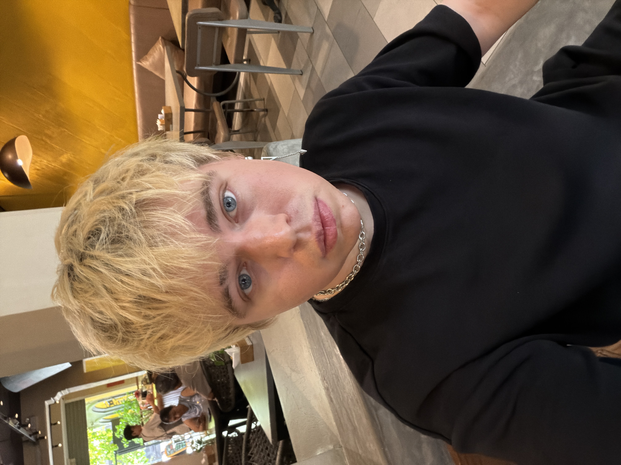 An individual with short, blond hair and blue eyes, wearing a black crew-neck top, a silver chain necklace, and a distinctive silver earring on the right ear, is positioned in the foreground, facing the camera. The setting is an indoor cafe or restaurant located in Thủ Đức, Vietnam.
In the left background, an open entrance reveals green foliage outdoors. Inside, a person in an apron is visible carrying items on a tray, and another individual is seated at a table. Dark, possibly wicker-style, tables and chairs are present in this area. Overhead, track lighting is visible on the ceiling.
The right background features an interior dining area with a textured, yellow-orange accent wall. A dark pendant light fixture hangs from the ceiling. Upholstered booth seating in a light brown tone lines the wall, accompanied by square tables and matching chairs. A wooden caddy holding condiments is observed on one table. The floor exhibits a checkered pattern of light and dark tiles.