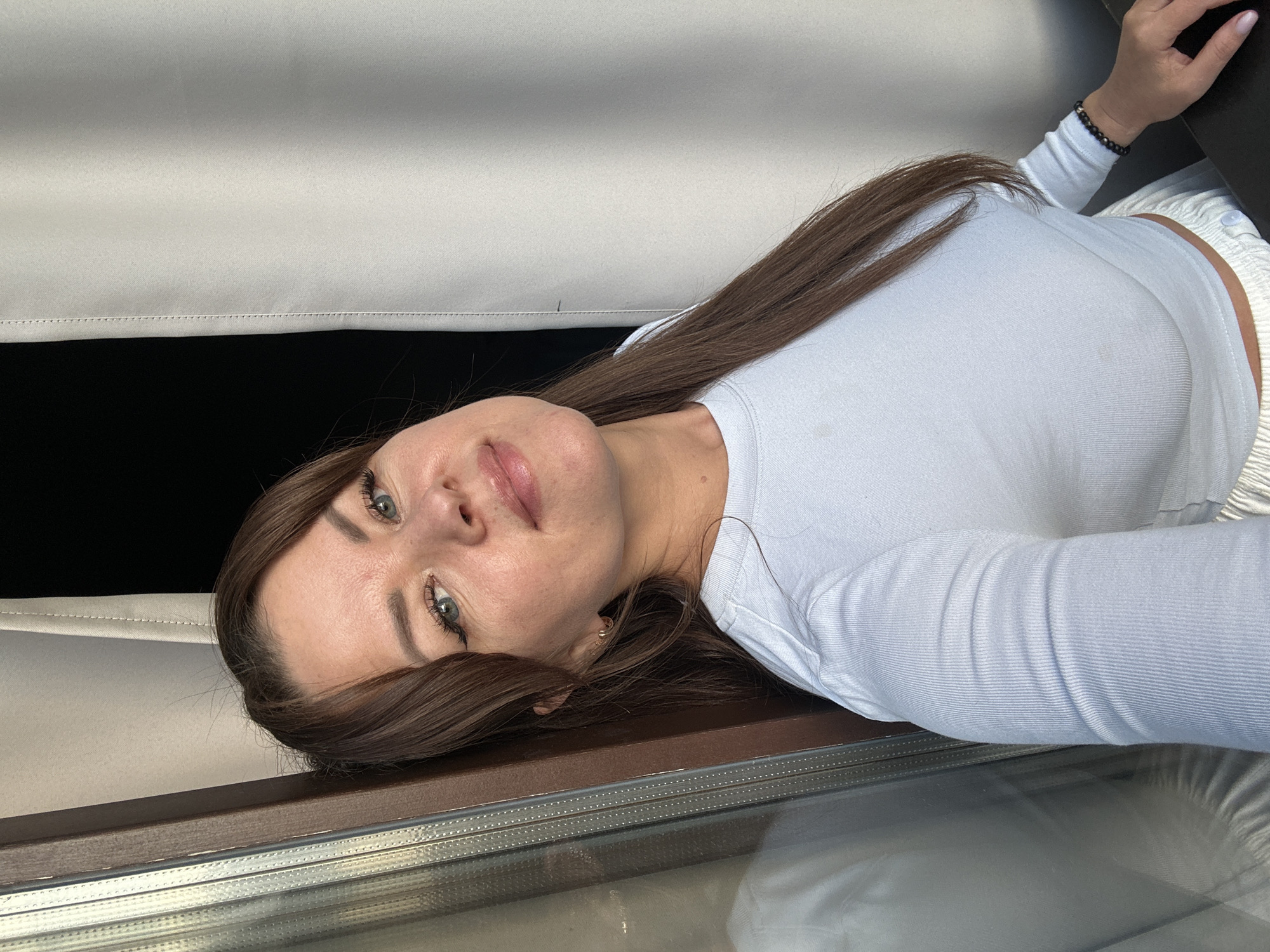 In a medium shot taken from a slightly low angle, a woman with long brown hair and light blue eyes is positioned in the foreground. She is wearing a white long-sleeved ribbed top and light-colored pants. The woman is leaning against a wooden frame of what appears to be a window or balcony door. To her right, a light-colored curtain is visible, partially obscuring a dark background. Her right hand rests on a dark surface, possibly a table or desk, and she is wearing a dark beaded bracelet on her right wrist. The overall scene suggests an indoor setting, possibly a room with a view, given the curtain and the window frame. The image does not provide specific indicators to confirm the location in the city of Chaikovsky, Russia.