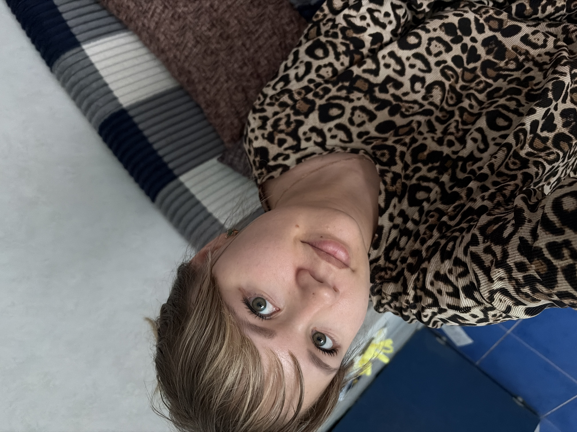 The image depicts a woman lying on her back. She is wearing a leopard-print top. The location is a room with a blue tiled surface and a striped blanket. The setting appears to be indoors, possibly in a private residence. There is no information that confirms the location is the city of Chaikovsky, Russia.