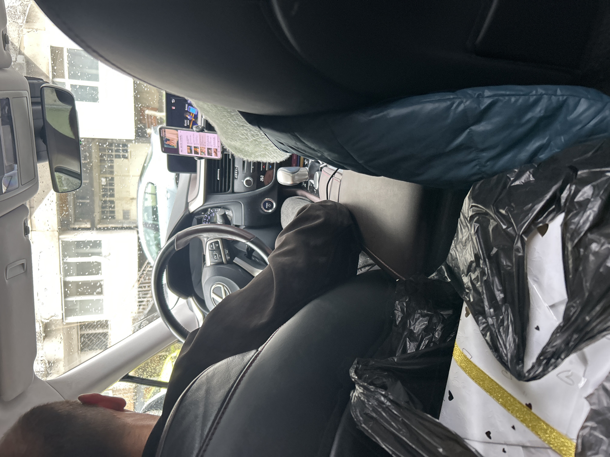 **Scene:** Interior of a car parked in Burgas, Bulgaria.
**Environment:** Cloudy, wet day outside. Buildings visible through the rain-streaked windows.
**People:** Partially visible is a person with short dark hair, possibly the driver.
**Objects:** Steering wheel, dashboard, phone mounted on the dashboard, rearview mirror, black garbage bag containing a white gift box with a gold ribbon.
**Actions:** Possible driving or parking activity.
**Interactions:** Uncertain.
**Additional Details:** The car appears cluttered.