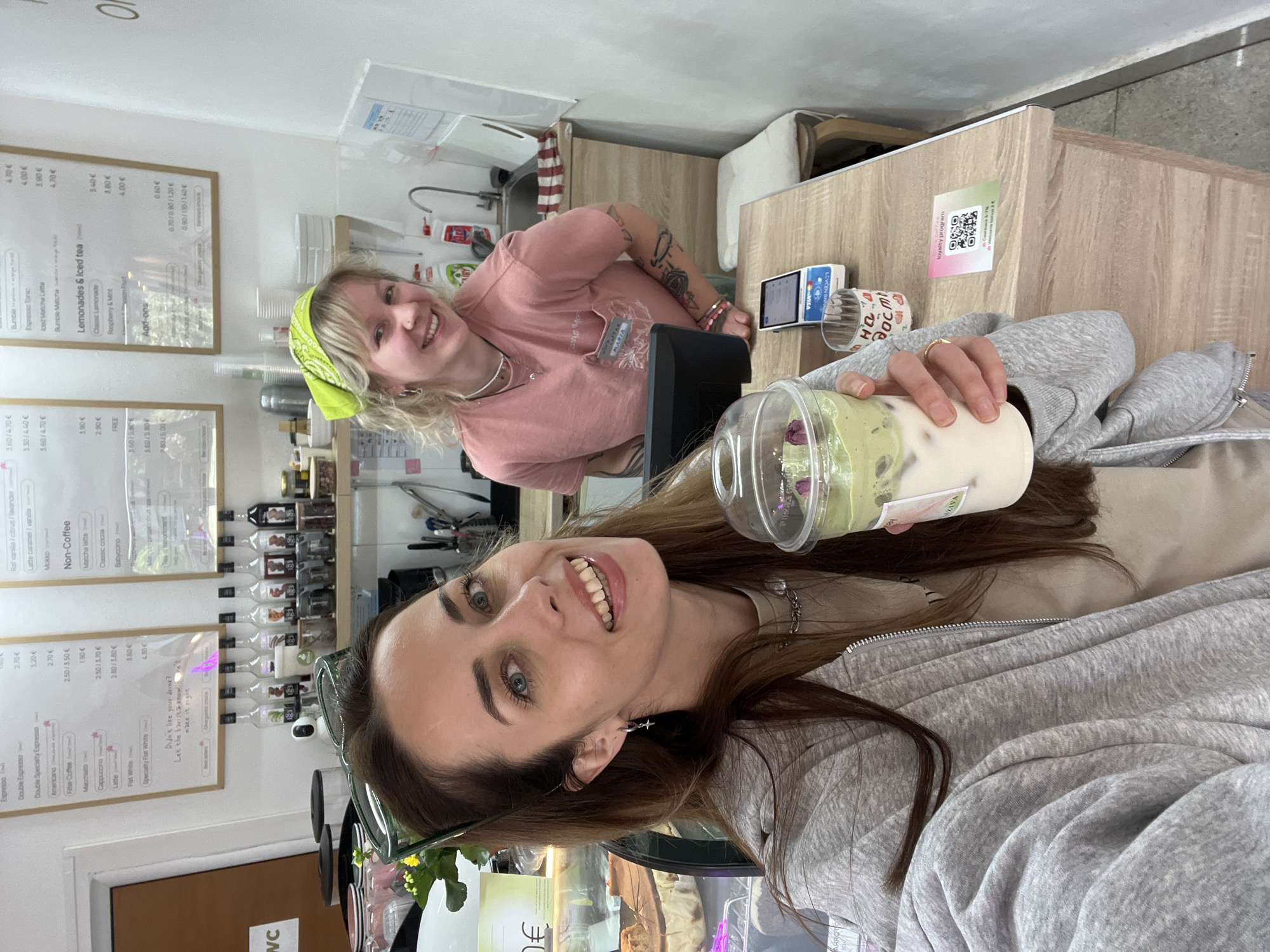 The image is a first-person perspective selfie, likely taken inside a café in Bar, Montenegro.
**People:**
* Two women are visible. The woman taking the photo is in the foreground, smiling and holding a clear plastic cup filled with a layered beverage (likely matcha and milk). She has brown hair, blue eyes, and wears a grey zip-up hoodie over a beige top.
* In the background, a second woman is behind the counter. She has blonde hair, wears a pink t-shirt, and also smiles at the camera. She has tattoos on her arms and wears a yellow bandana. She is identified as "Maria" via an ID badge.
**Objects:**
* The primary object is the beverage, consisting of layers, including what appear to be matcha and milk, adorned with pink flower petals. The beverage is in a clear plastic cup with a dome lid.
* Behind the counter, there is a point-of-sale system, a tip jar, and various café fixtures. There is a menu board on the wall listing various coffee and beverage options.
* There is a display case with baked goods visible in the lower-left foreground.
* A QR code for a loyalty program is visible on the counter.
**Actions/Interactions:**
* The woman in the foreground is holding up her beverage, likely showcasing it for the selfie.
* Both women are smiling, suggesting a friendly interaction or a positive experience at the café.
**Environment:**
* The location is a café with light walls, a wooden counter, and visible menu boards. The lighting is bright and even. A WC sign is visible on a door.
* The overall impression is that of a modern and inviting café environment in Bar, Montenegro.
