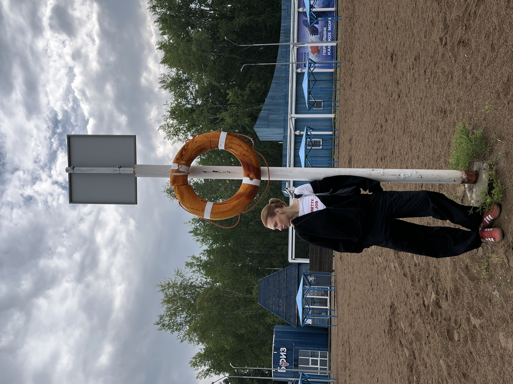 A woman stands next to a white pole with an orange life preserver attached in an outdoor sandy area. She is wearing a black hooded jacket, black pants, and red shoes. The background shows blue buildings with white canopies, trees, and a cloudy sky. The scene appears to be at a beach or recreational area in Chaikovsky, Russia, indicated by the text on the blue buildings.