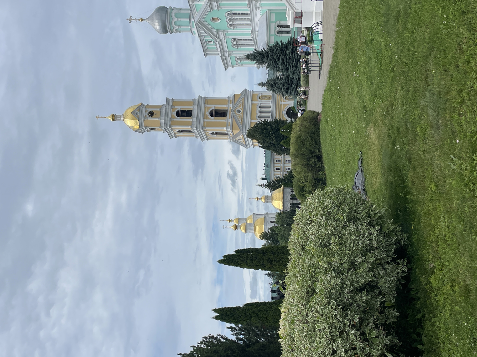 The image depicts a scenic view of the Seraphim-Diveyevo Monastery in Diveyevo, Russia. In the foreground, a lush green lawn with scattered small white flowers stretches across the frame, partially obscured by a large, variegated green and white bush on the left. The bush has a dense canopy and appears well-maintained. Behind the bush, a portion of a dark-colored object, possibly a tarp or bag, lies on the grass.
Dominating the mid-ground and background are several prominent architectural structures of the monastery. A tall, yellow bell tower with a golden dome stands prominently, featuring multiple levels with arched windows and a clock face. To its right is a building with a light blue facade and a silver dome, adorned with white decorative elements. Further to the left, several golden-domed churches are visible, some with white walls and intricate architectural details. Tall, dark green evergreen trees are strategically placed throughout the scene, adding height and contrast to the landscape.
A paved walkway or courtyard is visible in the mid-ground, where several people are scattered. Some are standing, others are seated on benches. They appear to be casually strolling or observing the surroundings. The sky above is overcast with a mix of gray and white clouds, suggesting a slightly diffused lighting condition. The overall atmosphere of the image is serene and picturesque, capturing the grandeur of the monastery and its peaceful grounds.