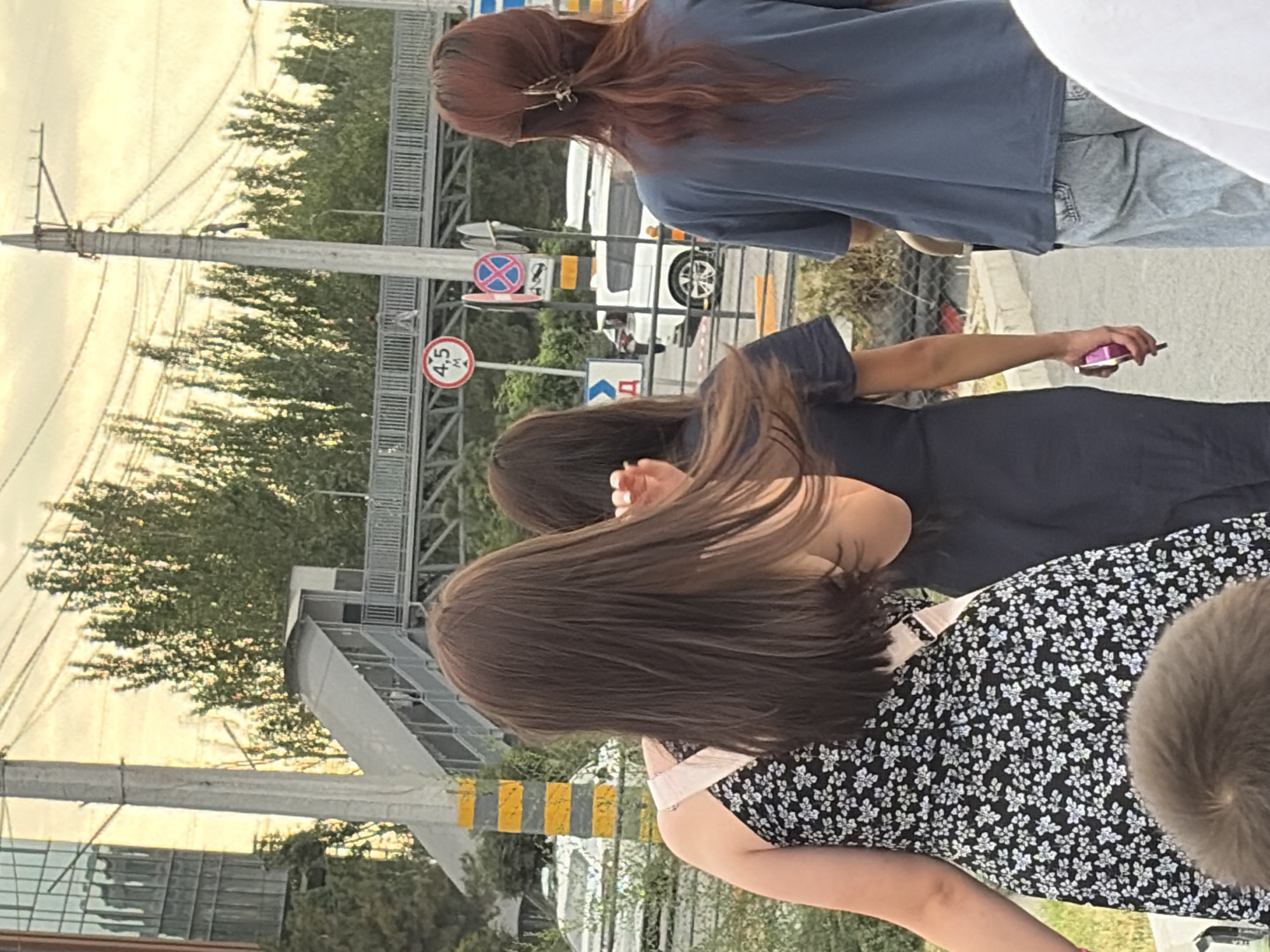 Three young women are walking away from the camera on a city street. The woman on the left is wearing a black and white floral dress and has long brown hair. The woman in the middle is wearing a dark blue dress and has long brown hair. The woman on the right is wearing a blue t-shirt and light-wash jeans, and has long reddish-brown hair pulled back in a ponytail with a clip. In the background, there is a pedestrian overpass with stairs, trees, a utility pole, and a white van. There are also traffic signs visible. The sky is overcast with a warm, yellowish hue.