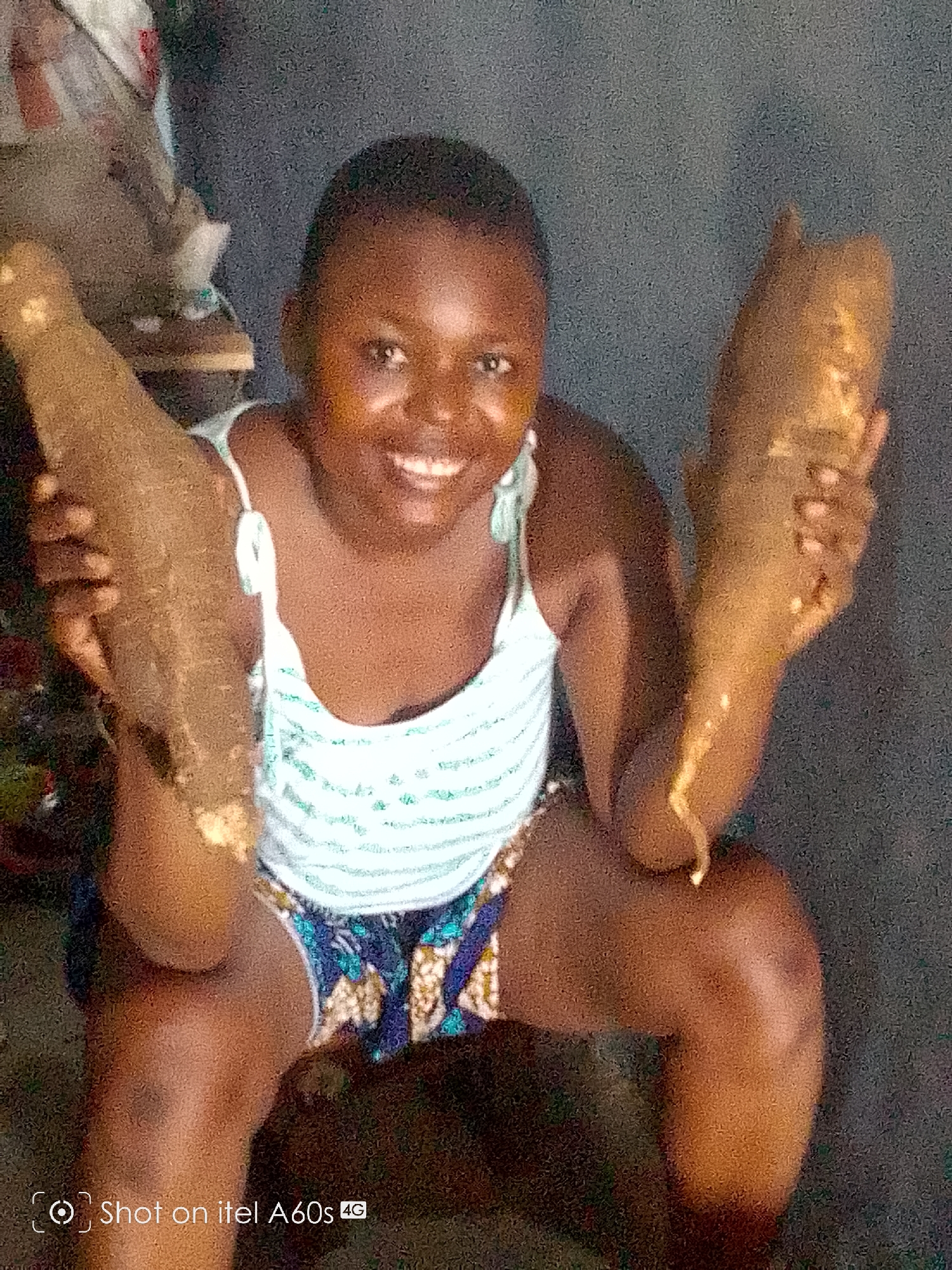 A smiling young woman squats indoors, holding two large, brown, root vegetables, possibly yams. She is wearing a white and light blue striped tank top and patterned shorts. The background is dimly lit and appears to be a simple interior space. The location is Ikeja, Nigeria, as indicated by the cultural context suggested by the woman's appearance and the food. The image includes a watermark indicating it was taken with an itel A60s phone.