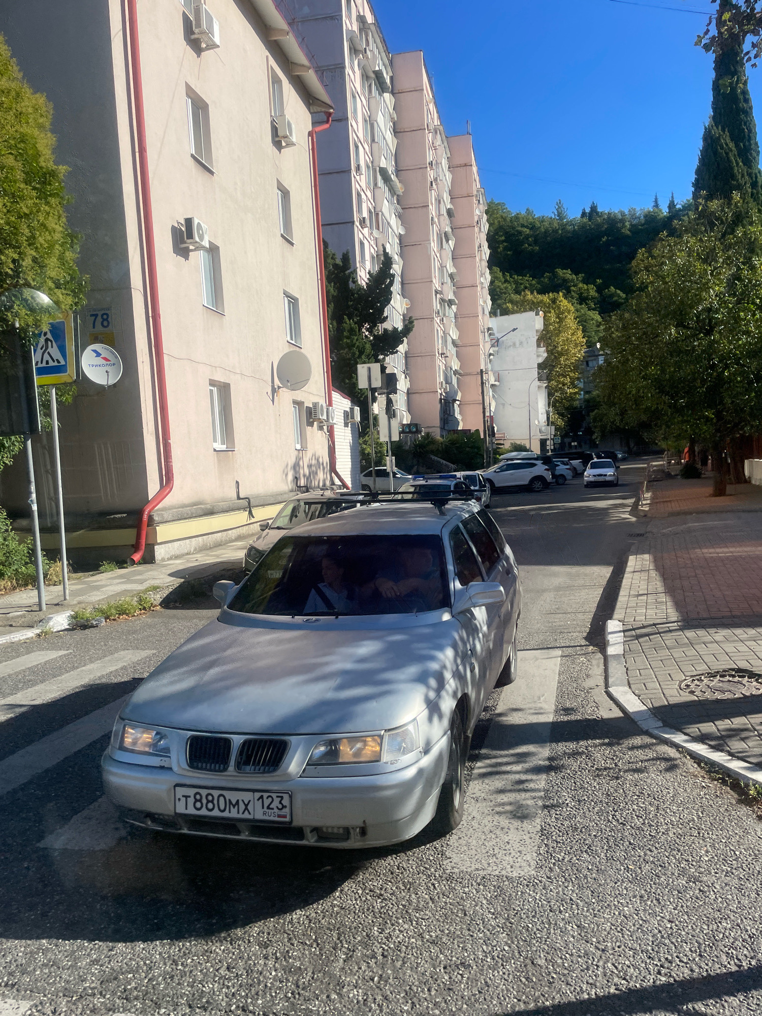 A silver station wagon is stopped at a pedestrian crossing on a street in Sochi, Russia. The car's license plate reads "T880MX 123 RUS". The vehicle is in the foreground, and in the background, there is a large, pale pink apartment building with multiple air conditioning units visible on its facade. A green tree with dense foliage is to the left of the building. Further down the street, other cars are visible, including a white SUV and a white sedan. The street is paved with asphalt and bordered by a sidewalk made of gray pavers. The sky is a clear, bright blue. A street sign with the number "78" and a satellite dish are mounted on the side of the apartment building. The scene suggests a sunny day in an urban environment.