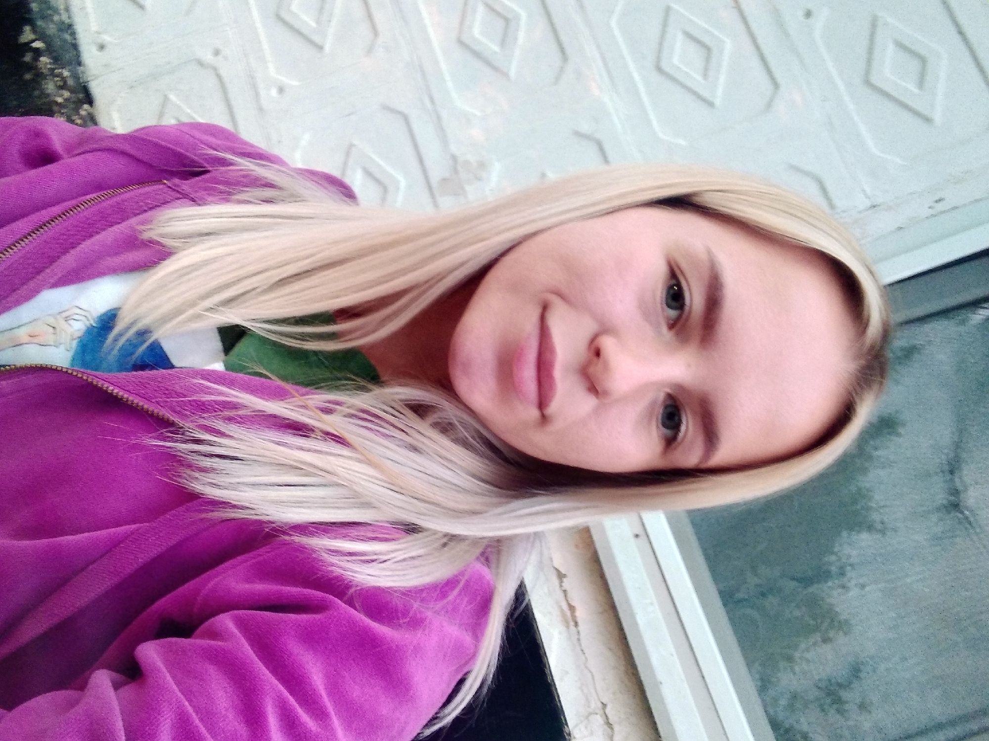 A young woman with blonde hair, wearing a purple zip-up jacket over a green shirt and a white t-shirt with a colorful graphic, is taking a selfie outdoors. She is positioned in the foreground, with a white textured wall on the left and a dark, possibly windowed surface on the right, likely in the city of Radisne, Ukraine. The lighting suggests it is daytime, possibly overcast.