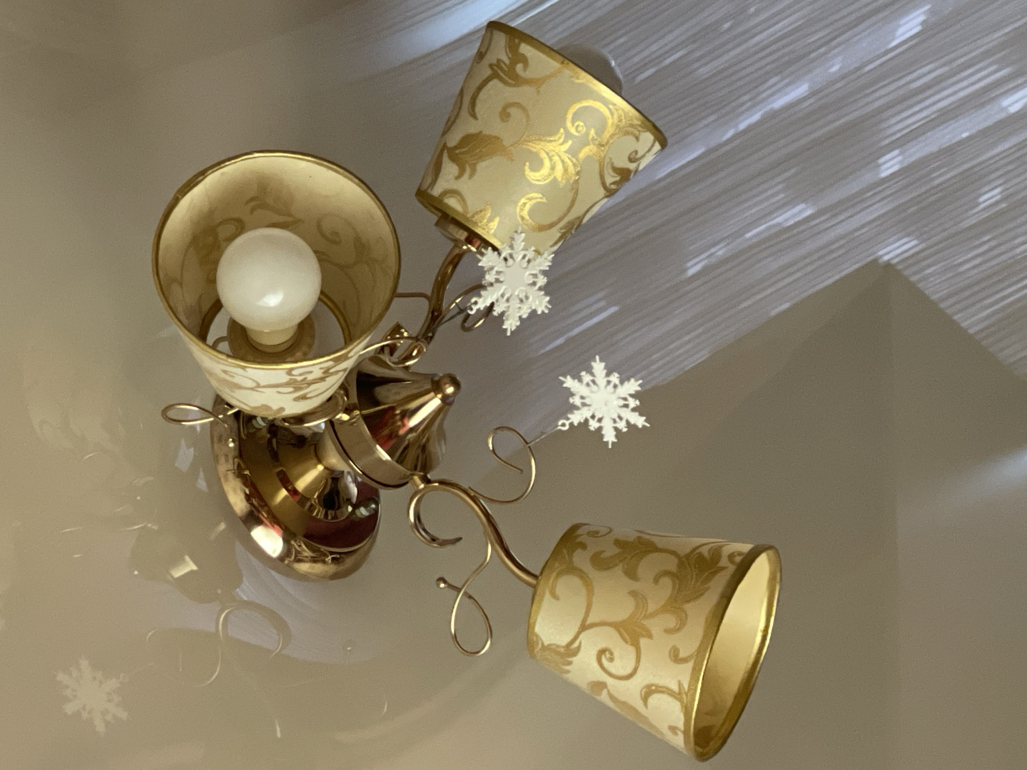 A ceiling light fixture with three lampshades is decorated with snowflake ornaments. The lampshades are conical, cream-colored with gold baroque patterns, and trimmed with gold. One shade has a lit bulb inside, and a small white snowflake is attached near its base. Another snowflake is suspended from the central fixture, and a third lies on the surface below, near the reflected image of the fixture. The ceiling has a ridged texture. The location is not discernible from the given image.