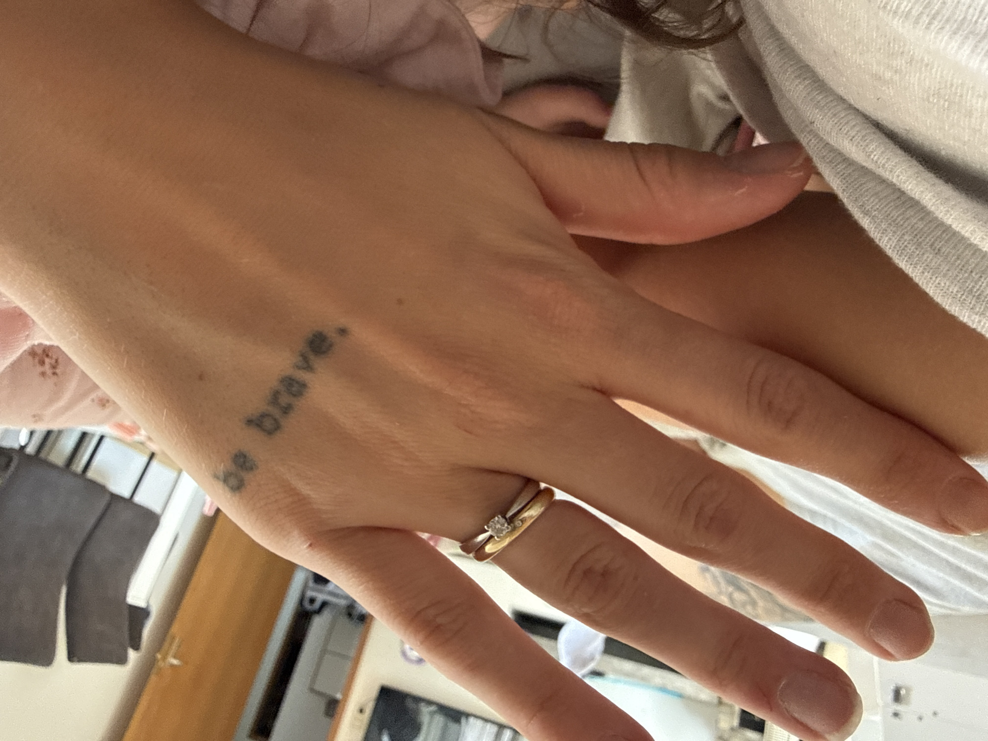 A close-up shot focuses on a person's left hand, adorned with two rings on the ring finger and a tattoo on the back of the hand. The tattoo, in black ink, reads "be brave." The hand is resting on a surface covered with a grey and white striped fabric, and partially over a pink fabric with a subtle pattern. In the background, out of focus, are two dark grey chairs, a wooden door, and what appears to be a white cabinet. The lighting suggests an indoor setting. The location is not specified.