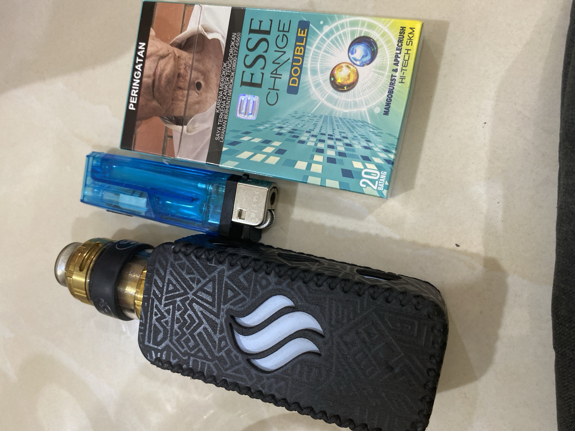 The image shows a pack of Esse Change Double cigarettes, a blue disposable lighter, and a vaping device on a light-colored surface, likely a tile floor. The cigarette pack features a warning image of a person with throat cancer. The vaping device has a black patterned case with a logo. The scene suggests a casual setting and depicts smoking and vaping-related items. There is no contextual information about location in Lhokseumawe, Indonesia, or people, actions, or interactions.