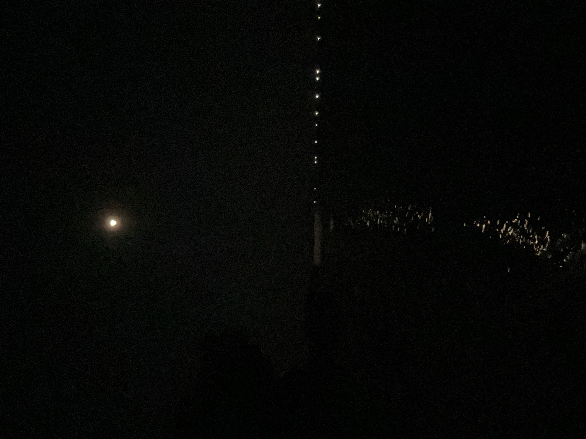 The image captures a night scene in Legonipah, Indonesia. The sky is dark, indicating it is nighttime. On the left side of the image, a bright, full moon is visible, casting a soft glow.
In the center of the image, a vertical line of small, bright lights is present, suggesting a string of lanterns or possibly lights from a tall structure. These lights are spaced evenly and descend from the top of the frame towards the bottom.
The right side of the image shows a dark area with scattered bright reflections. These reflections might be from water or wet surfaces illuminated by the lights in the scene. The ground appears uneven and possibly covered with vegetation.
There are no visible people or text in the image. The overall atmosphere is dark and mysterious, with the moon and the string of lights providing the main sources of illumination. The weather appears to be clear, as the moon is fully visible. The setting seems to be an outdoor environment, potentially near water or an area with a lot of foliage.