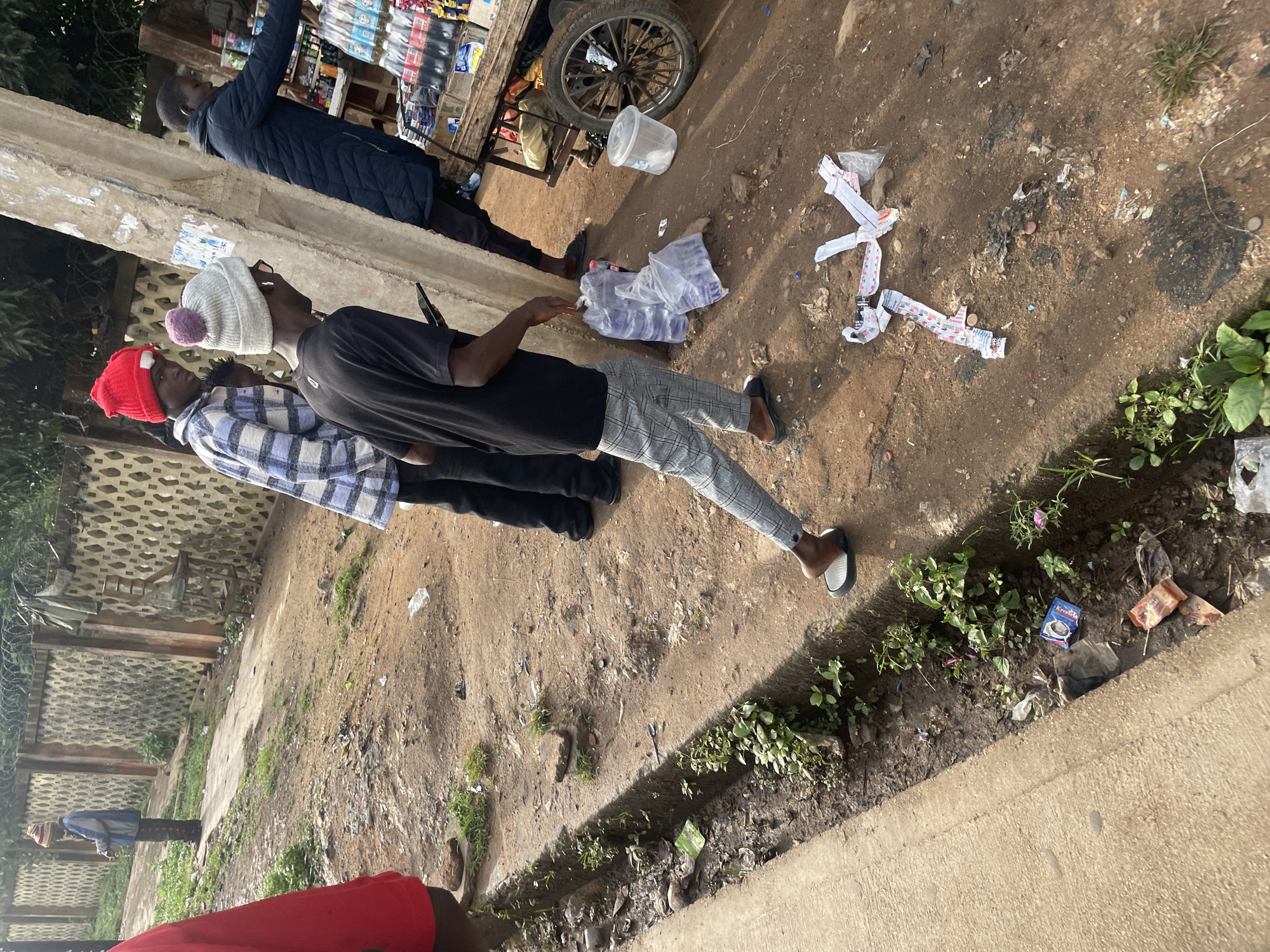 The image depicts an outdoor scene in Jos, Nigeria, characterized by a dirt and gravel ground with scattered debris. A concrete drainage channel runs along the left side of the frame, overgrown with sparse vegetation. Several individuals are present. In the foreground, a person wearing a black t-shirt, plaid trousers, and a grey beanie with a pom-pom is walking away from the camera. They are holding a mobile device in their right hand. To their left, partially visible, is another individual in a red beanie and a plaid shirt. Further back, a person in a dark blue jacket is interacting with a wooden cart laden with bottled beverages. The cart has a large spoked wheel. The background features a textured wall and a concrete pole. The overall impression is one of a street-side environment with evidence of commercial activity and urban detritus.