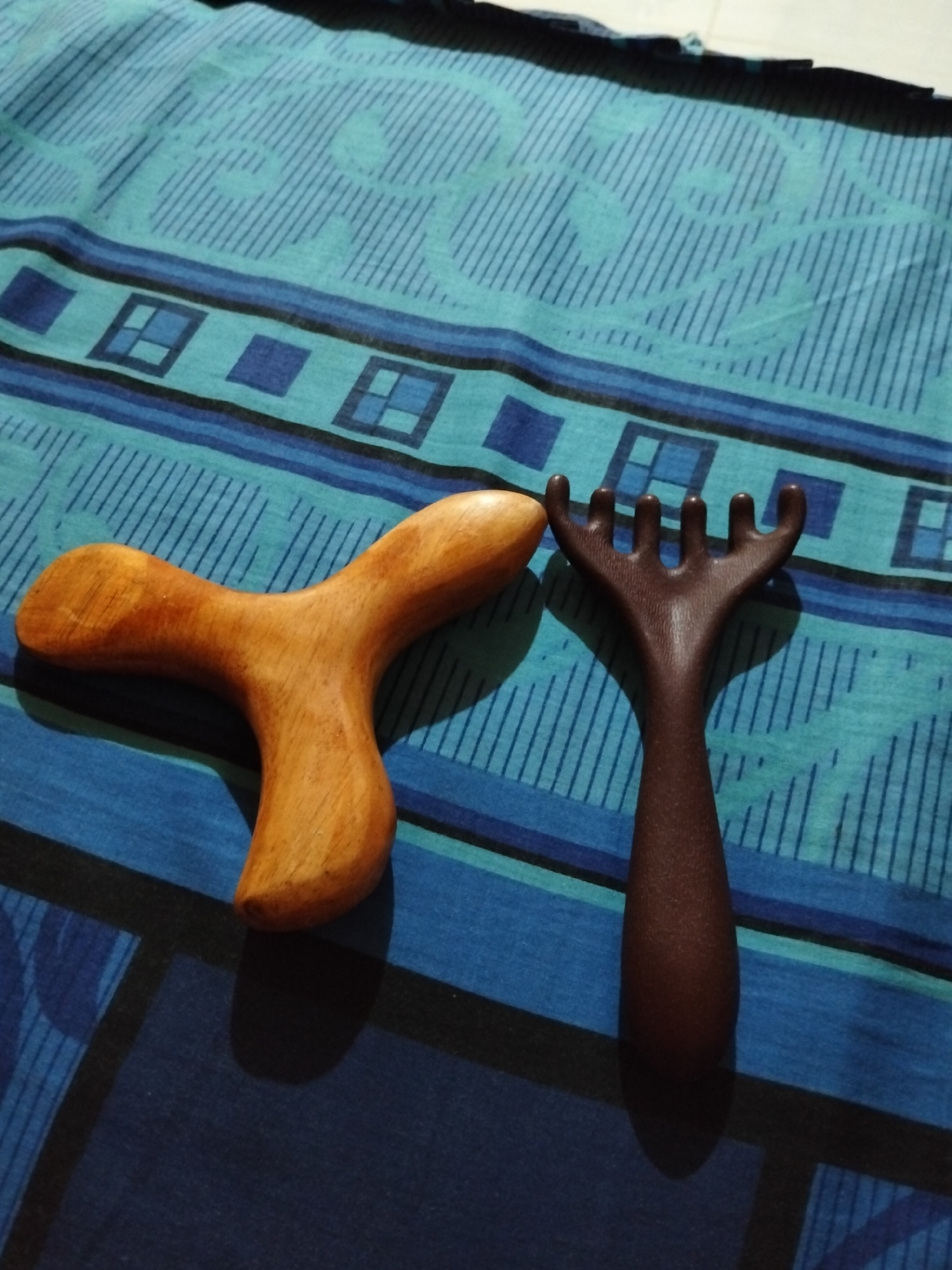 Two massage tools are placed on a blue and teal striped patterned fabric. The tool on the left is made of light brown wood and has a Y-shape with rounded ends. The tool on the right is dark brown, appears to be made of plastic, and resembles a hand with five fingers. The scene is lit from the top, casting shadows on the fabric. There is no indication of the location being Payagaboh, Indonesia.