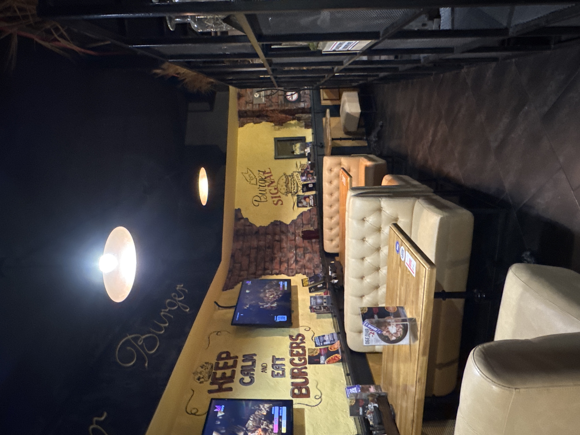 The image captures the interior of a casual dining establishment, likely a burger restaurant, viewed from a slightly elevated perspective. No people are visible.
The scene features a long, narrow dining area with seating arranged along both walls. On the left, a dark-colored wall (possibly black) contrasts with a bright, circular light fixture and cursive writing. The opposing wall is painted a vibrant yellow, adorned with brick-like painted sections and graphic murals. The ceiling has exposed metal beams, suggesting an industrial aesthetic.
The furniture includes comfortable-looking, light-colored upholstered booth seating with tufted details and matching chairs, paired with dark wooden tables. Several flat-screen televisions are mounted on the yellow wall, displaying what appears to be video games or entertainment content. Menus and flyers are placed on the tables.
No specific activity or event is taking place; the restaurant appears to be set up and awaiting customers. The time of day is not explicitly clear, but the reliance on artificial lighting suggests it could be evening or night.
Visible text includes:
* On the yellow wall: "Burger Signal" next to a burger graphic, and "KEEP CALM AND EAT BURGERS".
* On the dark wall: "Burger" written in a flowing script.
* On menus or flyers on the tables: "ОБЕДНА" (Russian for "Lunch" or "Dining") and "BBQ 390".
* On the televisions: Some unreadable text, but "NY3" or "NYT" is faintly discernible, possibly part of a game interface.
The presence of Russian text "ОБЕДНА" on the menus confirms the location context of Russia.