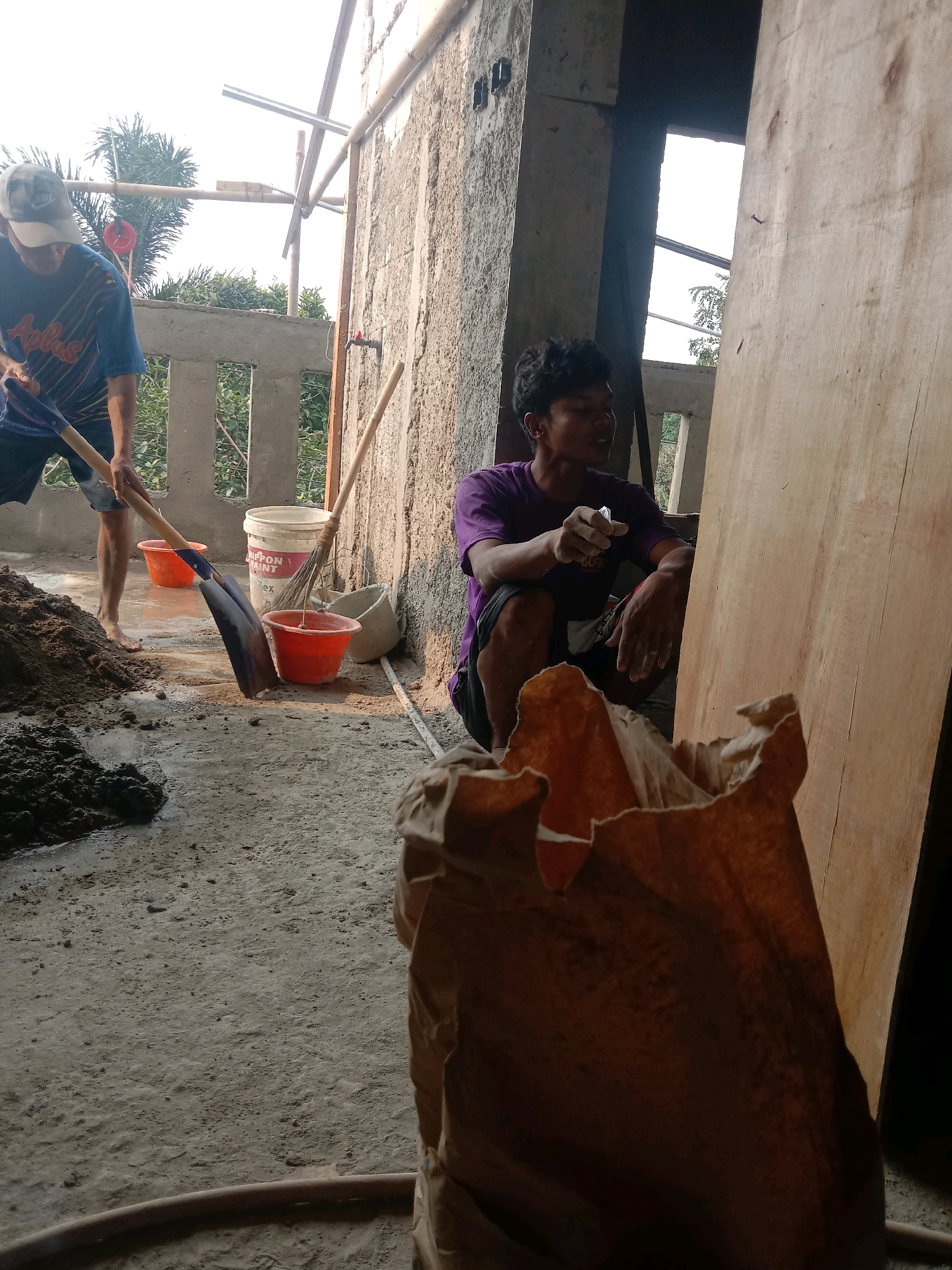 Two men are working on a construction site in Tajurhalang, Indonesia. One man, wearing a blue and yellow striped t-shirt and a white baseball cap, is standing and using a shovel to mix cement. A pile of cement mix is next to him. Nearby, a red bucket and a white paint bucket are visible. Another man, wearing a purple t-shirt, is squatting and appears to be gesturing with his hands, possibly instructing or discussing the work. He is positioned in front of a large sheet of plywood. The environment suggests an unfinished building with concrete walls, scaffolding, and a concrete floor. The time of day appears to be afternoon, with sunlight casting shadows.