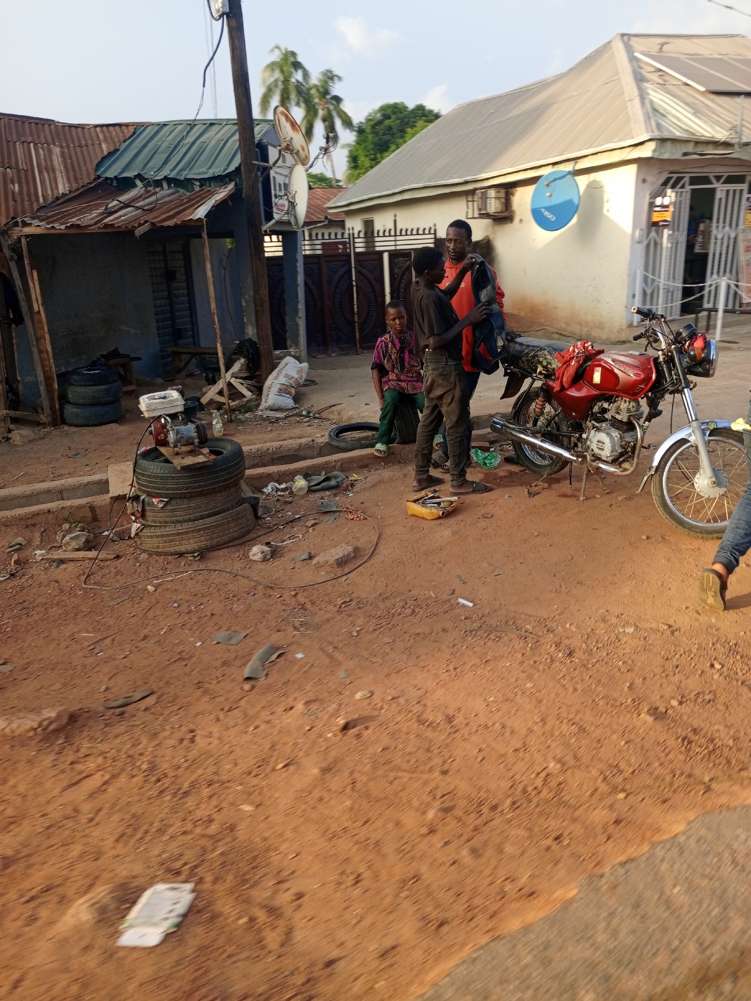 The scene is in Keffi, Nigeria. The environment is dry and dusty with a red, clay-like soil. A red motorcycle is parked on the roadside, near a group of three people. Two appear to be older, while one is a child. They are standing around a collection of tools and tires, suggesting they may be repairing the motorcycle or related machinery. A small generator is placed on top of a stack of tires. Buildings in the background are a mix of corrugated iron structures and more permanent buildings with satellite dishes. A utility pole with electrical wiring is also visible. The overall impression is a somewhat cluttered, informal roadside setting with individuals engaged in mechanical work.