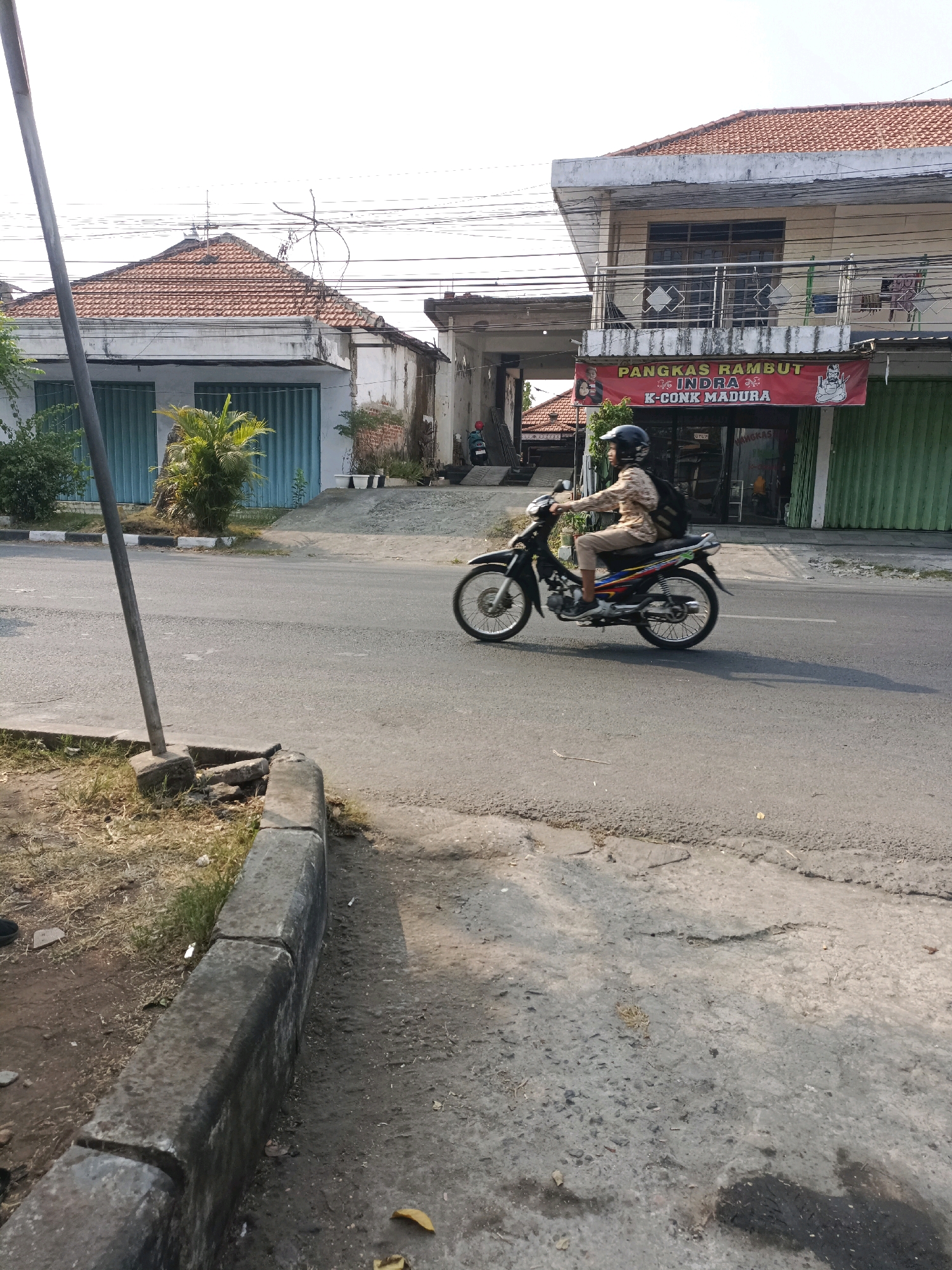 A person wearing a helmet and backpack rides a motorcycle on a paved road in front of shops. The motorcycle is black with colorful accents and the rider is wearing a patterned shirt and light-colored pants. In the background, there are buildings with red tile roofs and green shutters. A banner above one of the shops reads "Pangkas Rambut Indra K-Conk Madura." The scene appears to be in an urban or semi-urban area with power lines visible overhead. The road has some cracks and patches, and a concrete curb runs along the side.
