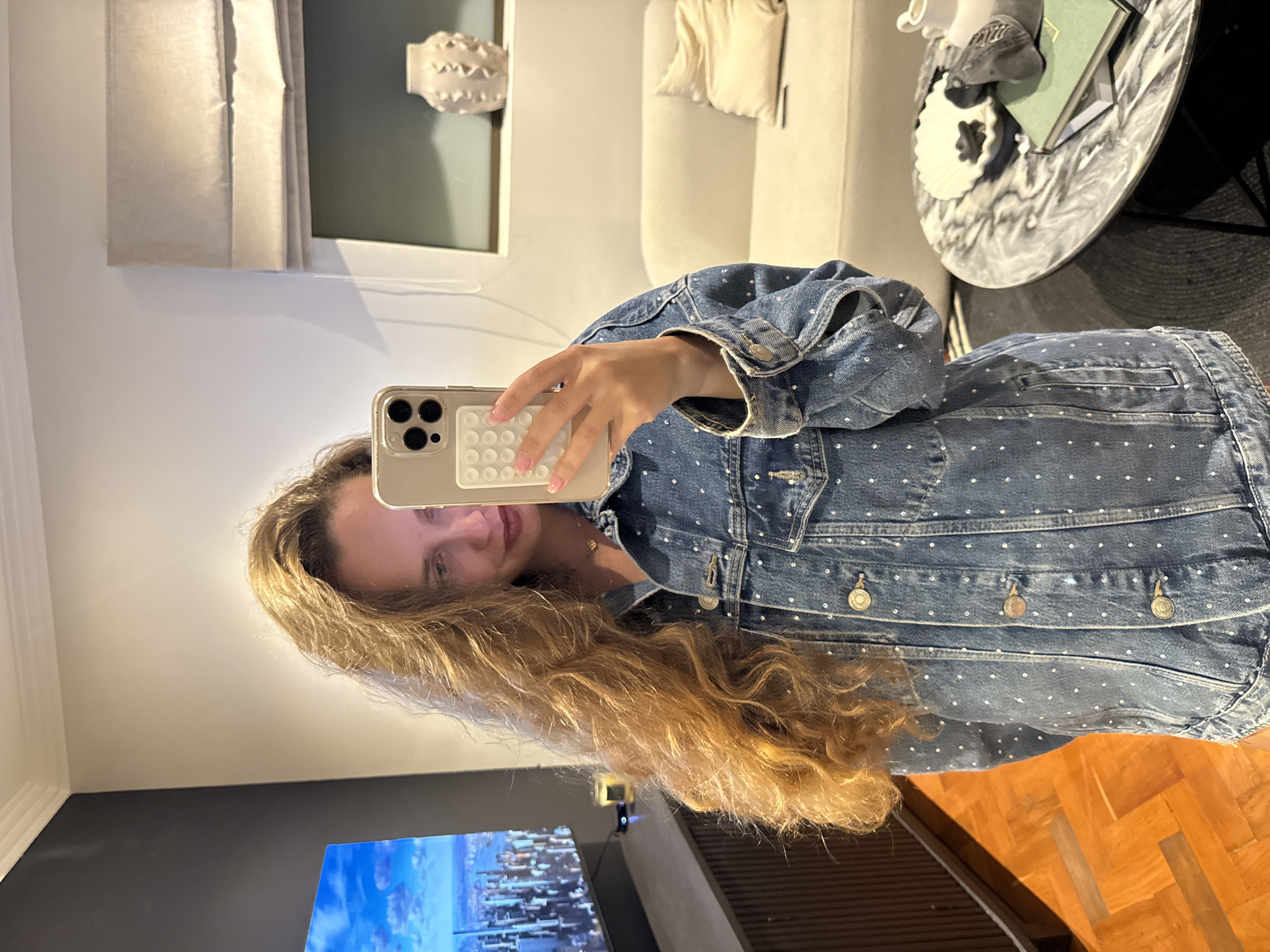 A young woman with long, blonde hair is taking a selfie. She is wearing a blue denim jacket with white polka dots. She is holding a gold iPhone with a white pop-it case. The background shows a living room with a white sofa, a round marble coffee table, and a television. The television screen displays an image of Cape Town, South Africa. There is a window or artwork visible in the background. The woman appears to be inside her home. The flooring is wooden parquet.