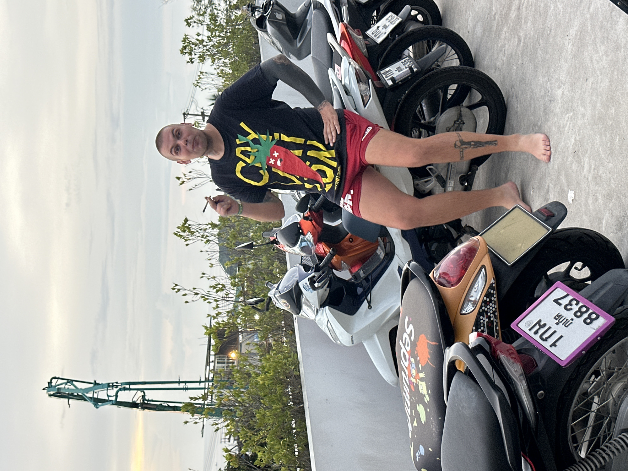In Karon, Thailand, a man with a shaved head and multiple tattoos, including one on his left arm and a crucifixion tattoo on his right shin, sits on a white motorcycle. He is wearing a black t-shirt with a yellow and red chili pepper graphic and red shorts, and he is holding a cigarette in his right hand. He is barefoot. Several other motorcycles are parked behind him, including a black one and an orange one. In the background, there are green bushes, a white wall, and a tall, green structure that appears to be a construction crane. The sky is overcast.
