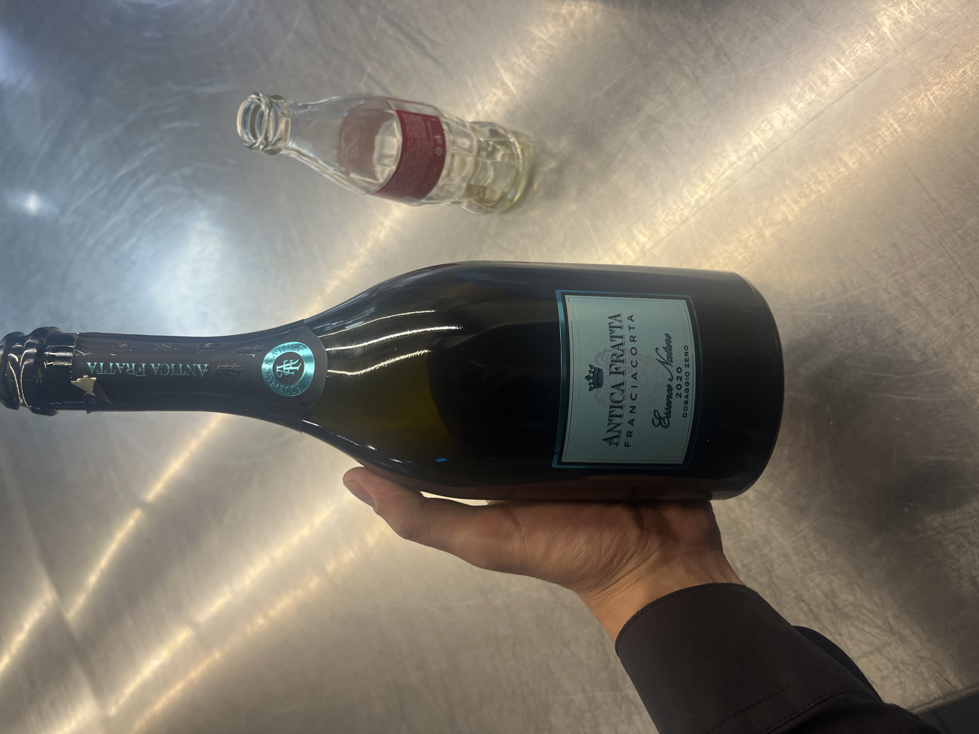A human hand, dressed in a dark long-sleeved shirt, holds a dark-colored bottle of sparkling wine horizontally over a highly reflective, metallic surface. The bottle features a light blue label that prominently displays the text "ANTICA FRATTA FRANCIACORTA" at the top, with "Essence Nature" and "2020 DOSAGGIO ZERO" visible below. A decorative crest or logo is also present on the label. The bottle's neck is sealed with a dark foil wrapper, which shows minor creasing.
In the background, lying on its side on the same metallic surface, is an empty, clear glass Coca-Cola bottle, recognizable by its iconic shape and a visible red label. The surface, likely a stainless steel countertop, reflects bright streaks of light, indicating a well-lit indoor environment. This setting suggests a professional kitchen, bar, or restaurant. The image captures a paused moment, perhaps before the wine is opened or served. No specific time of day or emotional cues are discernible, and the scene is entirely indoors. The overall context suggests an establishment in Sofia, Bulgaria.
