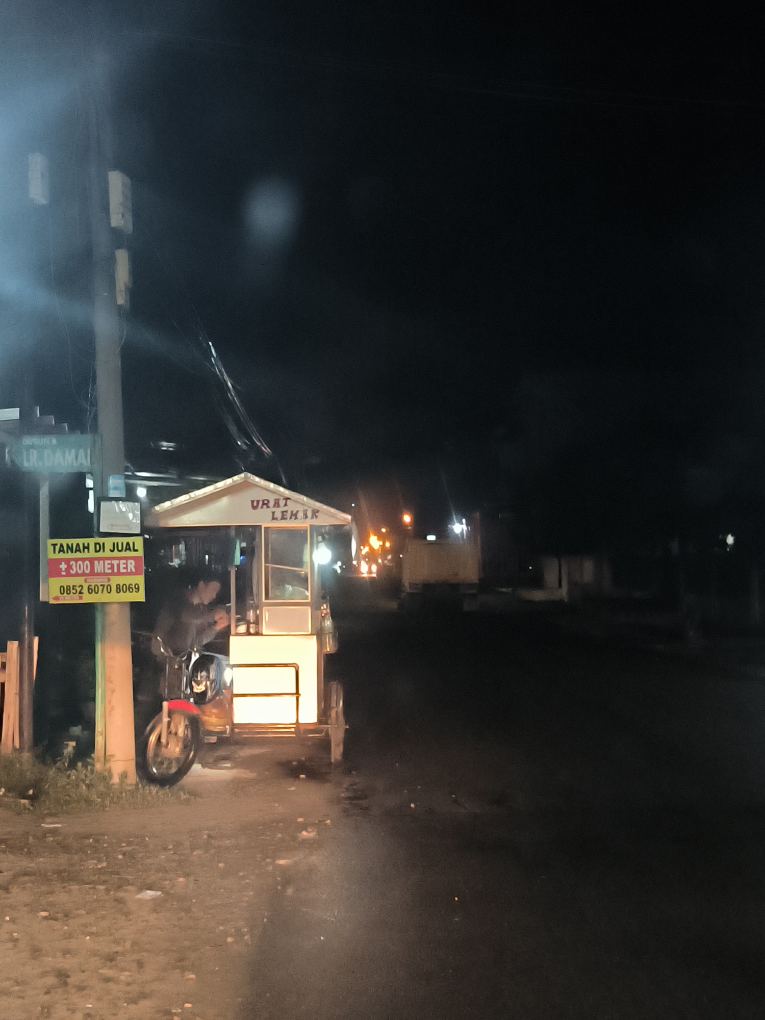 This nighttime photograph captures a street scene in Lhokseumawe, Indonesia, characterized by darkness punctuated by artificial light sources. The primary subject is a brightly lit mobile food stall built onto the rear of a red motorcycle, parked on the left side of a dimly lit, unpaved road. A person is visible sitting on the motorcycle, seemingly operating the stall.
To the left of the food stall, a utility pole stands with multiple signs. A street sign at the top reads "DUSUN I" and "LR. DAMAI". Below it, a yellow rectangular sign with red lettering advertises "TANAH DI JUAL" (Land for Sale), stating "± 300 METER" and providing a contact number: "HUBUNGI: 0852 6070 8069". On the roof of the food stall, the words "URAT LEHER" are clearly visible, likely indicating the name or specialty of the stall.
The surrounding environment is mostly shrouded in darkness, with some distant lights visible further down the road and in the background, suggesting other activity or structures. The ground beneath the stall and pole appears to be dirt or gravel, possibly damp. The image conveys a common sight of a street food vendor operating in the evening hours.