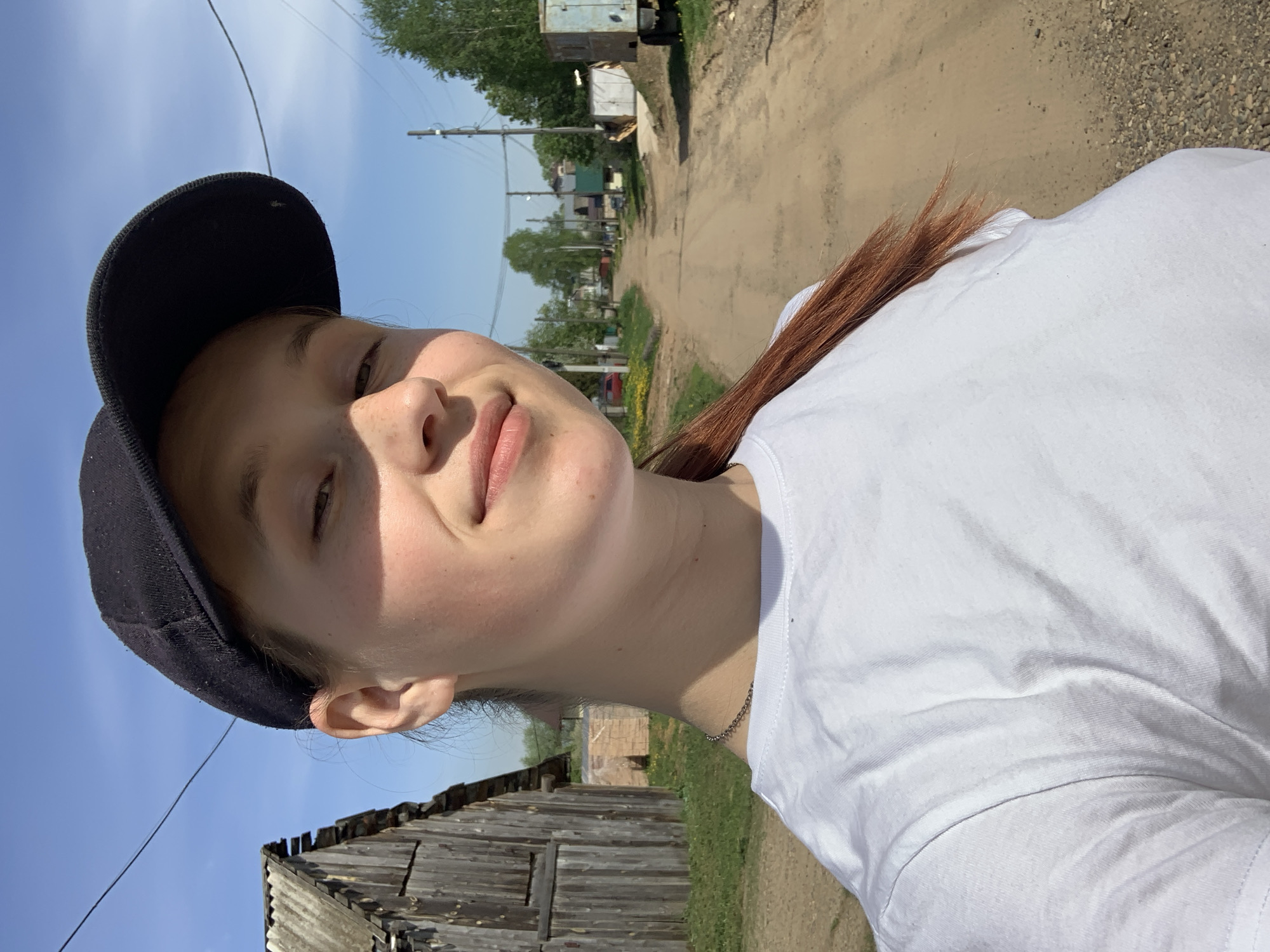 A young, fair-skinned woman wearing a white t-shirt and a black baseball cap takes a selfie outdoors. She is looking directly at the camera with a slight smile, and her red hair is visible behind her shoulder.
The background suggests a rural setting within the city of Фоки, Russia. An unpaved dirt road stretches into the distance. There is a weathered wooden structure to the left of the frame, suggesting older or less developed architecture. Vegetation, including trees and patches of grass, lines the road. Power lines are visible overhead, adding to the environment.