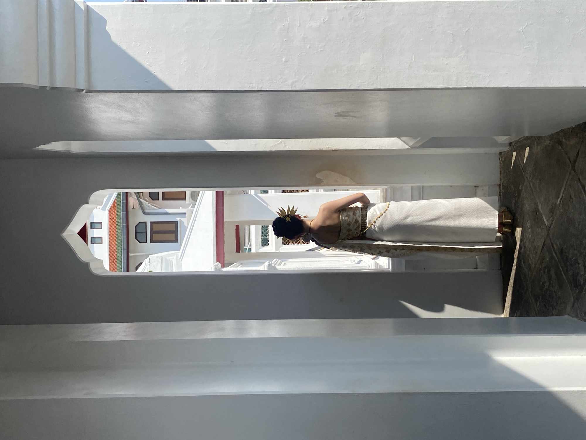 A person wearing traditional Thai clothing stands in a white building. The person's back is to the viewer, and their head is adorned with a golden headpiece. The location is likely a temple in Bangkok, Thailand, given the architectural style of the building in the background. The image is framed by the arched opening of the structure the person is standing in, which has multiple white horizontal bars. The exterior building visible through the arch is white with a red and green roof. The steps outside the archway are gray.