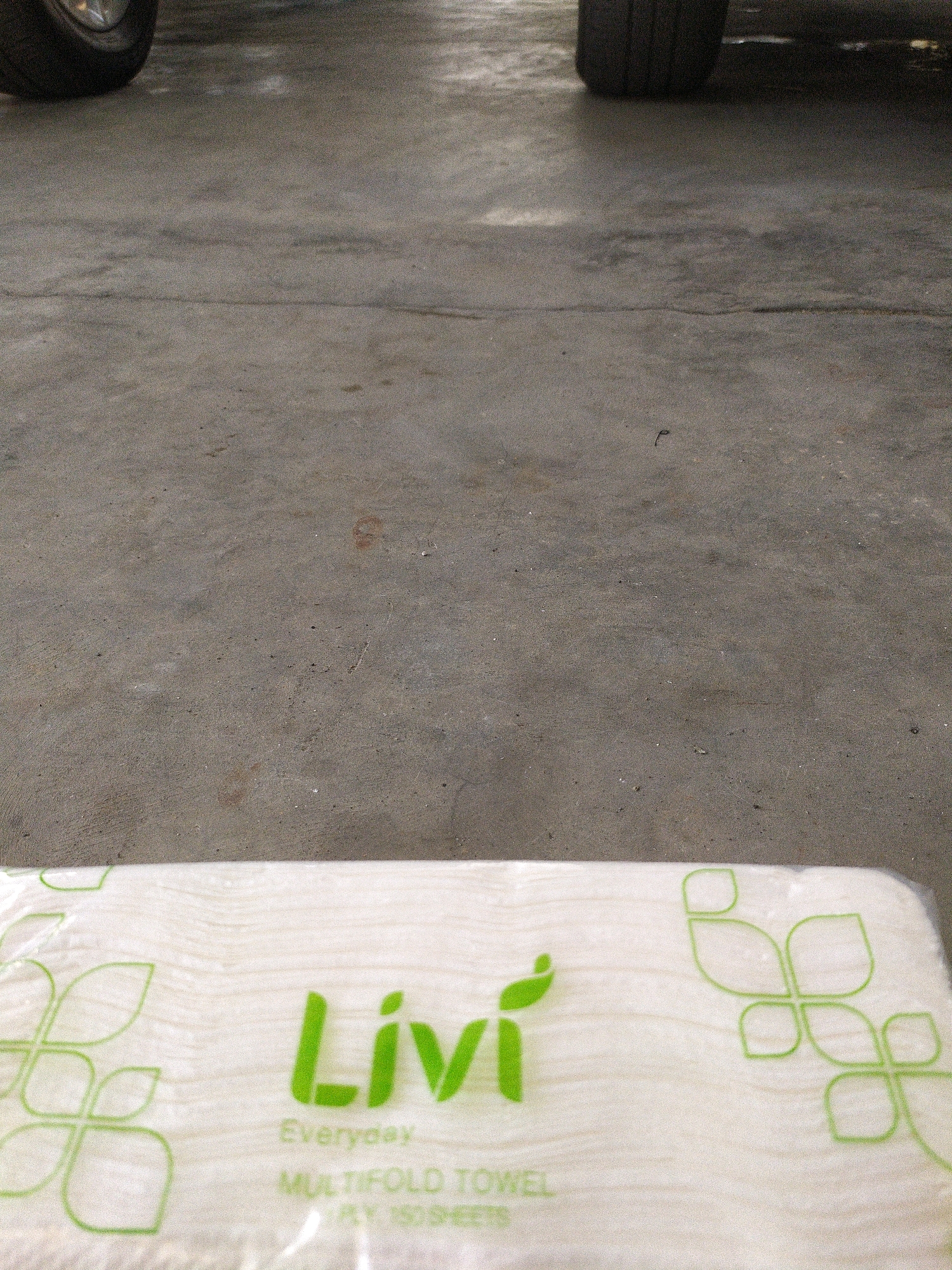 The image shows a close-up of a package of Livi brand Multifold Towels on a concrete surface. The package is partially wrapped in clear plastic and features green branding with the Livi logo and the text "Everyday" and "MULTIFOLD TOWEL". The concrete surface appears to be outdoors, possibly a driveway or sidewalk, with tire tracks visible in the upper portion of the image. The background is out of focus, showing what appears to be a car's tire. There are no people present in the image. The setting is a mundane, everyday environment.
