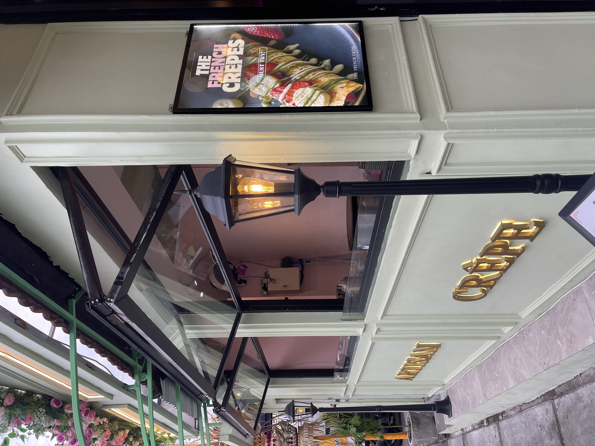 This is a medium shot, eye-level, outdoor, full-color photograph of a creperie facade in North Kuta, Indonesia. The building features a light green exterior with decorative molding and gold lettering that reads "ARTISAN CRÊPE." The creperie has an open-air counter with a glass window and an awning. A black lamppost with a lit bulb stands in front of the counter. To the right of the entrance, a backlit sign displays a picture of a strawberry and banana crepe with the text "THE FRENCH CREPES MUST TRY!". Flowers are visible above the awning, and a small tree is planted in a gray pot to the left of the entrance. The street in front of the building is paved with gray stones. There are no people visible in the photograph.