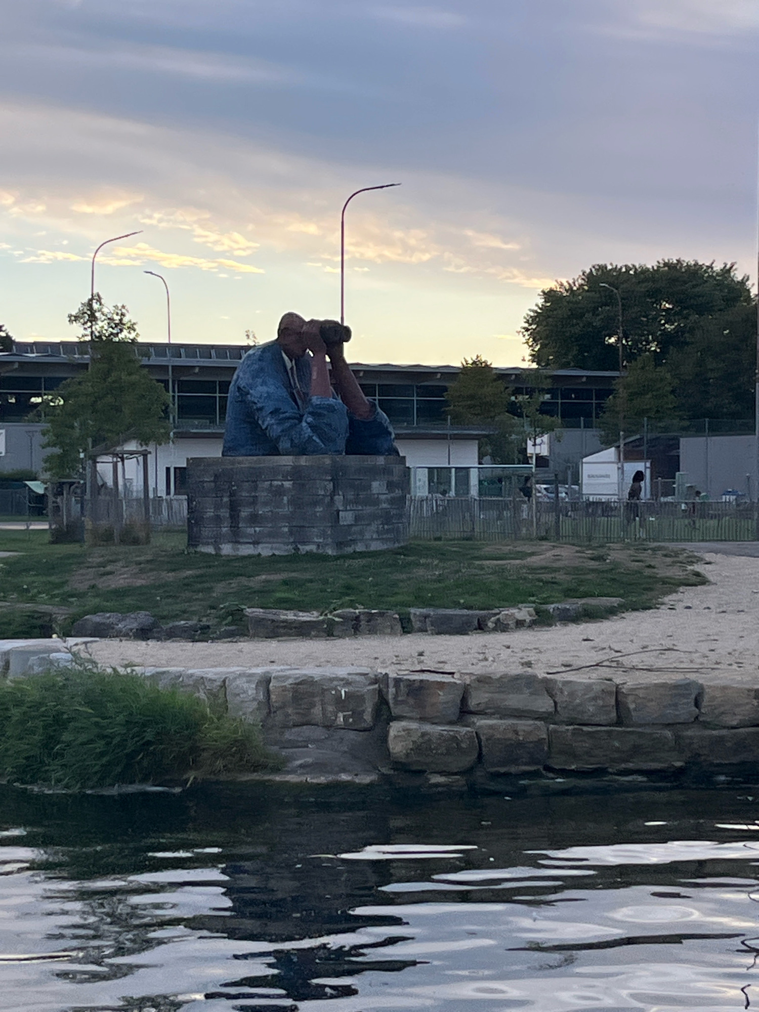 The image shows a large statue of a man sitting and looking through binoculars, positioned on a stone pedestal in an outdoor public space in Yverdon-les-Bains, Switzerland. The statue appears to be made of a rough, textured material, possibly concrete or a similar composite, and is depicted wearing a blue jacket. The background includes trees, a modern building with large windows, streetlights, and a fence. In the foreground, there is water with reflections, and a retaining wall made of large stones separates the water from the grassy area where the statue is located. The sky is overcast with some patches of lighter clouds, suggesting it might be late afternoon or early evening. There are no other prominent human figures visible interacting with the statue or the immediate environment.