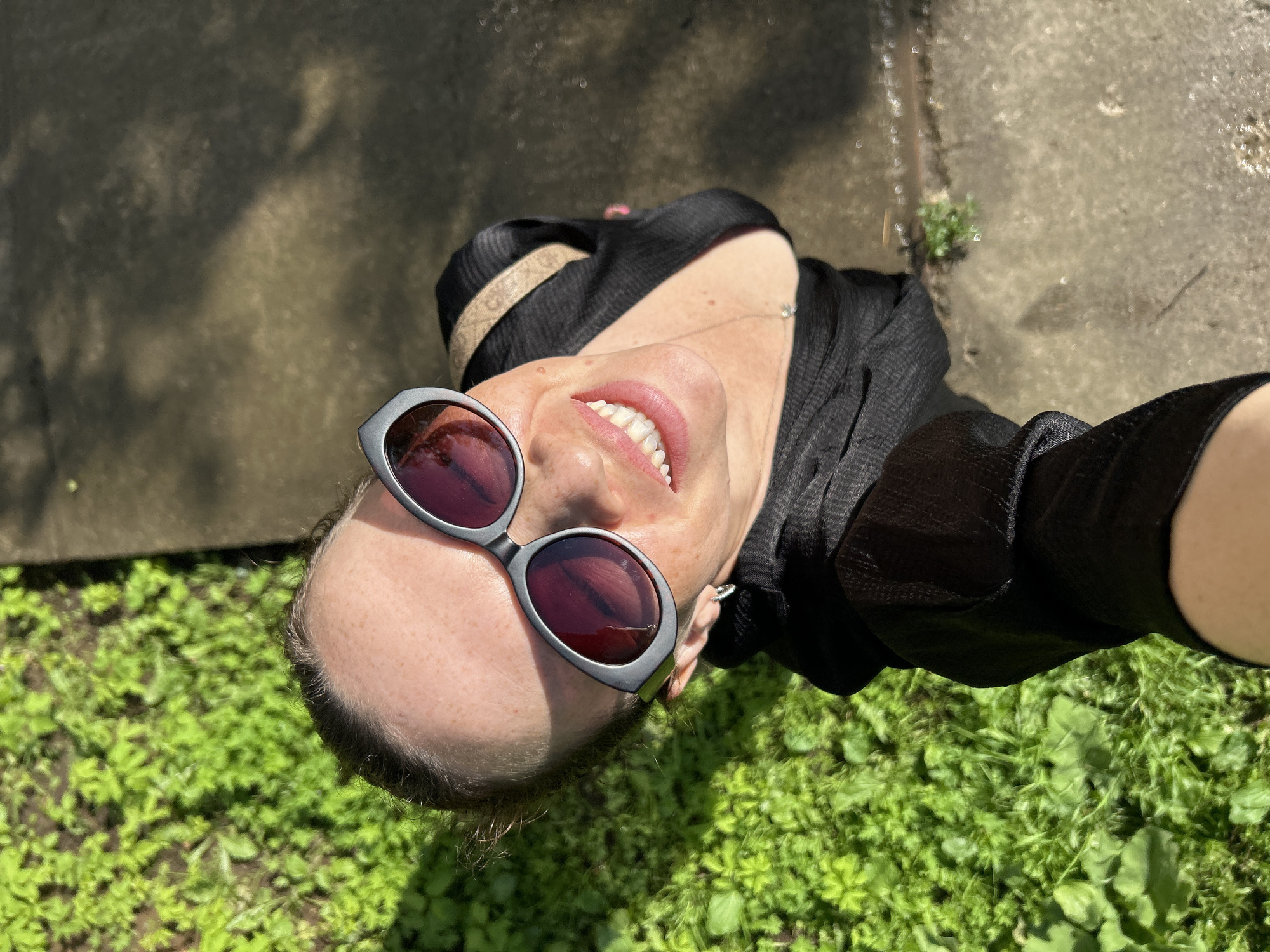A high-angle, eye-level shot captures a woman in Chaikovsky, Russia, during daylight. She is looking up at the camera and smiling broadly, her mouth open to reveal her teeth. Her hair is pulled back, and she is wearing large, grey-rimmed sunglasses with dark lenses. She is also wearing a black top with a low neckline, and a beige strap is visible across her shoulder. The background is divided into two distinct areas: on the left, lush green grass with sunlight dappling its surface, and on the right, a wet, grey paved surface that appears to be a sidewalk or path, reflecting the sunlight in scattered, bright spots. The overall impression is that of a casual outdoor snapshot.