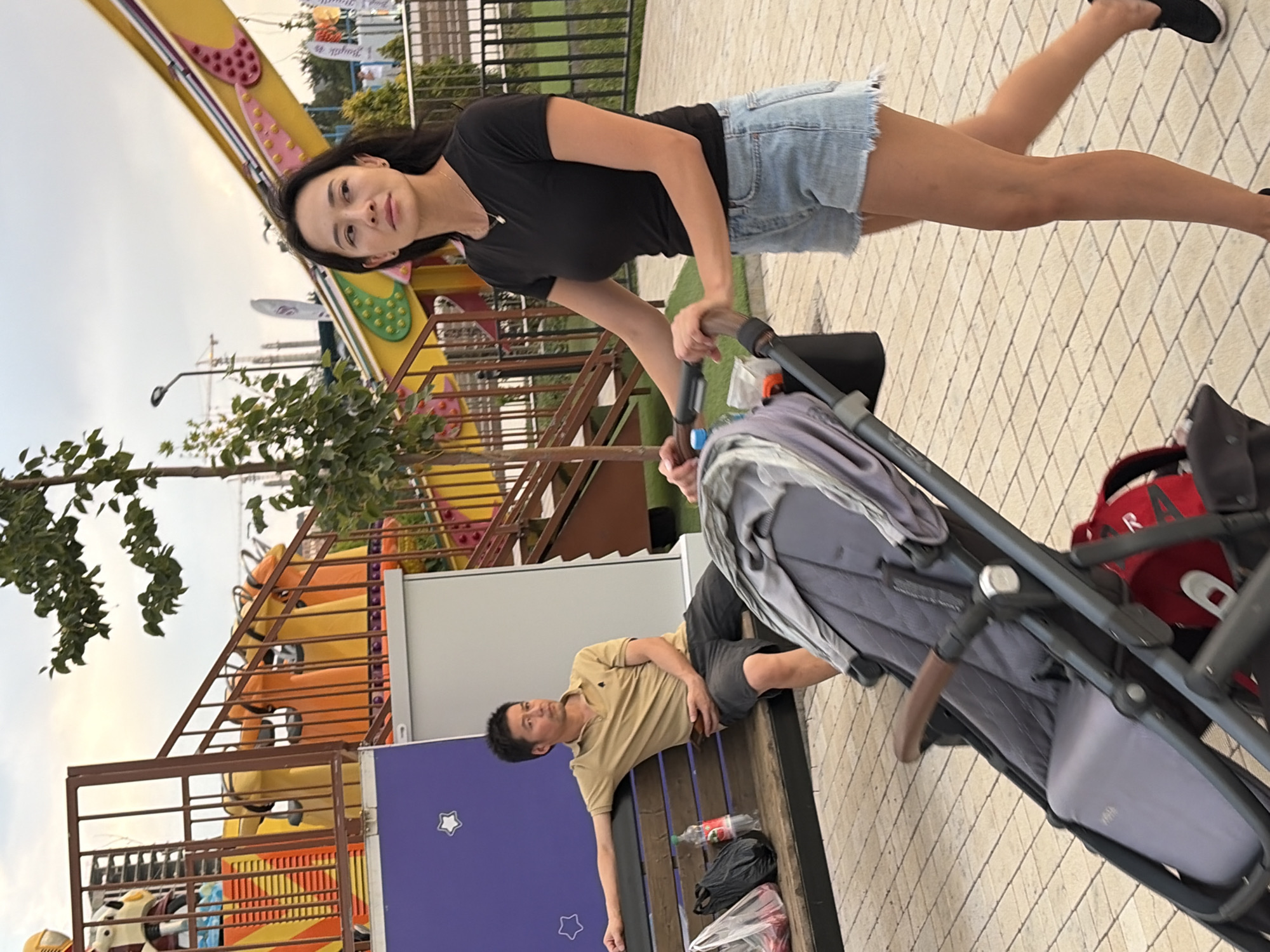 An Asian man in a tan polo shirt and dark shorts sits on a wooden bench to the left of the frame. A plastic bottle with a red label and a black plastic bag are on the bench beside him. To the right, an Asian woman with dark hair, wearing a black t-shirt and denim shorts, walks to the right, pushing a grey stroller. The stroller is in the foreground. In the background, there is a playground ride with yellow and orange structures, and a large yellow amusement park attraction with a purple stripe. The scene takes place on a paved walkway under an overcast sky.