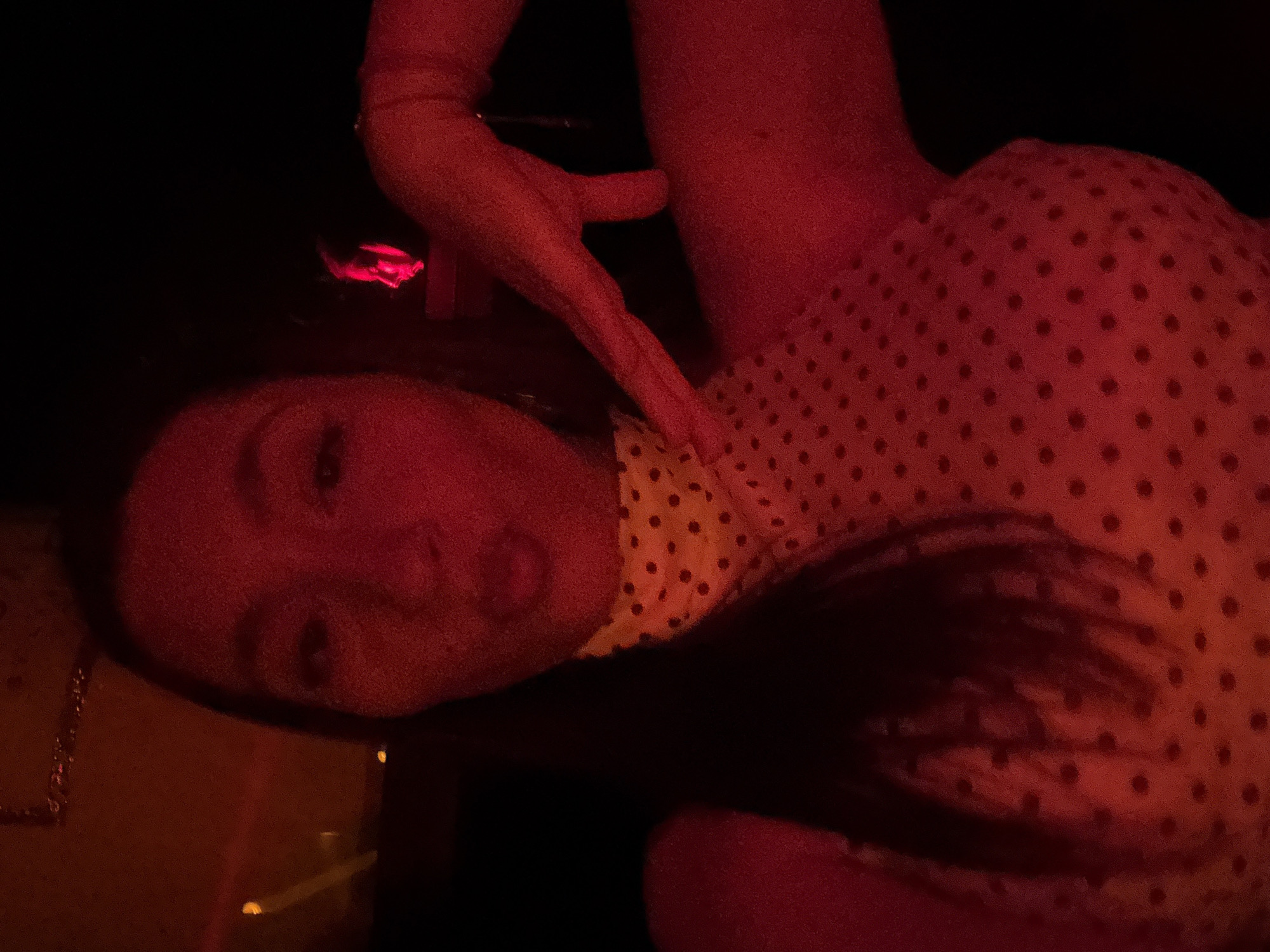 A woman is captured in a selfie taken under red lighting, likely in a nightclub or similar dimly lit entertainment venue in Sofia, Bulgaria. She is wearing a yellow top with black polka dots. Her lips are pursed, and her right hand is extended towards her left shoulder, with her index finger pointing slightly upward. In the background, a blurred reflection of another person, also illuminated by red light, can be seen. The overall ambiance is dark with a strong red hue.