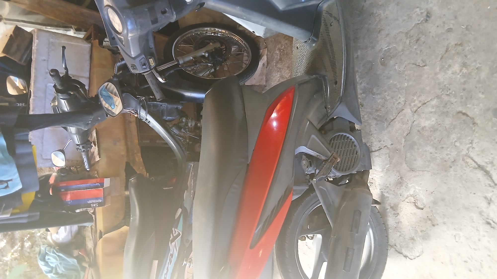 This media file captures a cluttered scene in what appears to be a garage or workshop, located in Kecamatan Pati, Indonesia. The main subjects are several motorbikes or scooters. A prominent dark grey scooter with vibrant red side panels is positioned in the foreground, showcasing its seat, handlebars, and a portion of its exhaust system. Another dark grey or black scooter is visible towards the top of the frame, revealing its handlebars, front wheel with spokes, and part of its fairing. A third scooter, partially obscured, with a blue and black color scheme, can be seen in the bottom left.
The activity depicted is the static storage or parking of these vehicles. The setting is characterized by a concrete floor with visible cracks, and various miscellaneous items are scattered around, including what looks like a large white foam block and blue fabric. Cardboard boxes are stacked on the left side; one box clearly displays the text "SNG" and "SMS". The scene is brightly illuminated by strong natural light, suggesting it is daytime, possibly with sunlight entering from an unseen opening, causing some areas to be overexposed. No people are explicitly visible, and no specific event or activity, such as repair or riding, is taking place.