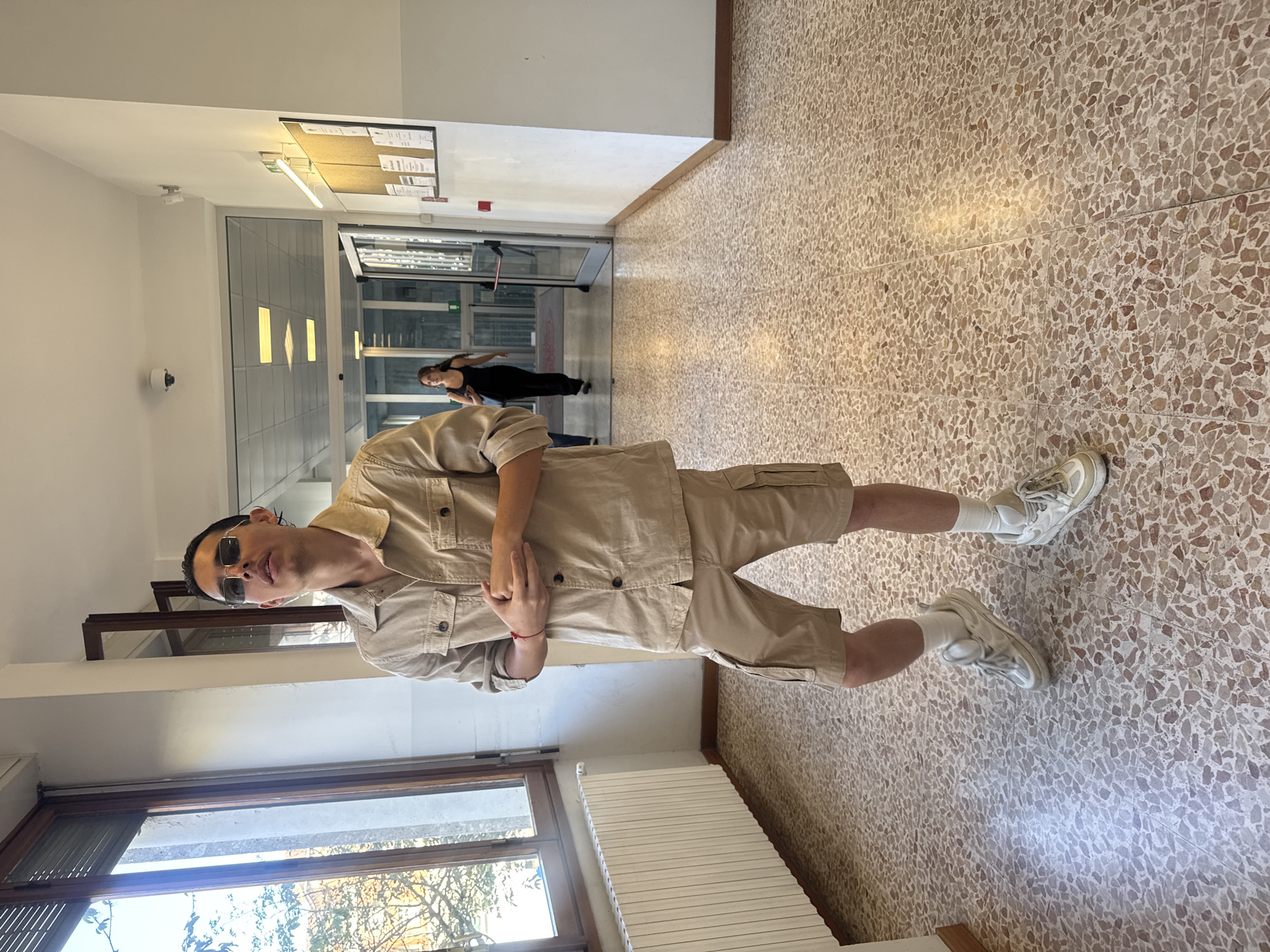 The image depicts a full-body portrait of a young man posing in an indoor hallway, likely in a building located in Milan, Italy. The man, with dark, slicked-back hair and dark sunglasses, stands confidently, looking directly at the camera with a relaxed expression. He is dressed in a monochromatic beige outfit, comprising a long-sleeved shirt with front pockets and matching cargo shorts. He completes his look with white socks and light-colored sneakers. A subtle red bracelet is visible on his right wrist, and his arms are crossed in front of him.
The hallway features light-colored walls and a speckled terrazzo floor, composed of various shades of brown and beige tiles. Natural daylight streams in from a large window on the left side of the frame, revealing glimpses of tree branches outside. A white panel radiator is installed beneath the window. In the background, a woman wearing a black top and dark pants is seen walking away, heading towards a set of glass or automatic doors. Further along the hallway, a bulletin board covered with various papers is visible on the wall, alongside a mounted security camera. The bright illumination suggests it is daytime. No specific text on the bulletin board or other objects is legible. The setting appears to be a public or institutional environment such as a school or office building.