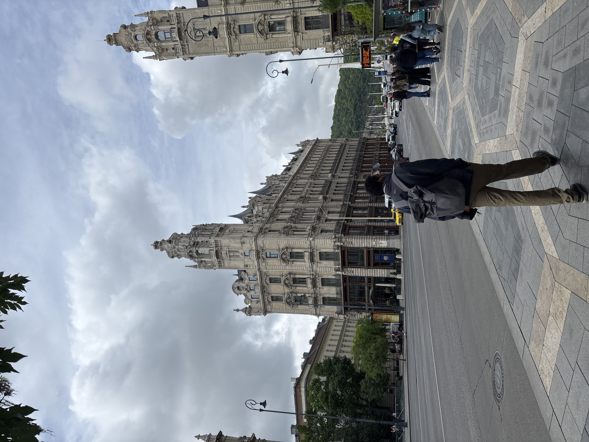 This is an eye-level outdoor shot in Budapest, Hungary. The buildings are of classic European design, high and embellished with architectural details. Pedestrians are walking on the right. Some trees can be seen on the bottom left. The sky is covered with gray clouds. The street is gray. A person with a backpack is walking in the right foreground. A street light can be seen in the bottom left.