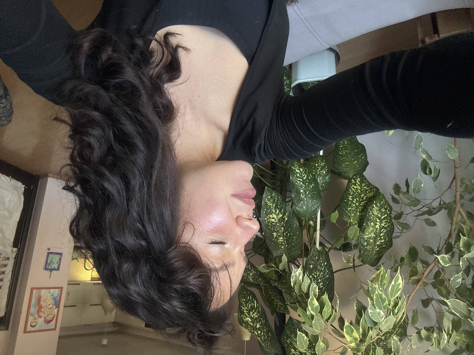 A woman with dark, curly hair is wearing a black long-sleeved shirt. Her eyes are closed, and she is positioned in front of multiple plants with variegated green and yellow leaves. In the background, a domestic interior is visible, including framed artwork on a pink-colored wall, suggesting a room inside a house or apartment. There is no clear evidence indicating the location as Burgas, Bulgaria.