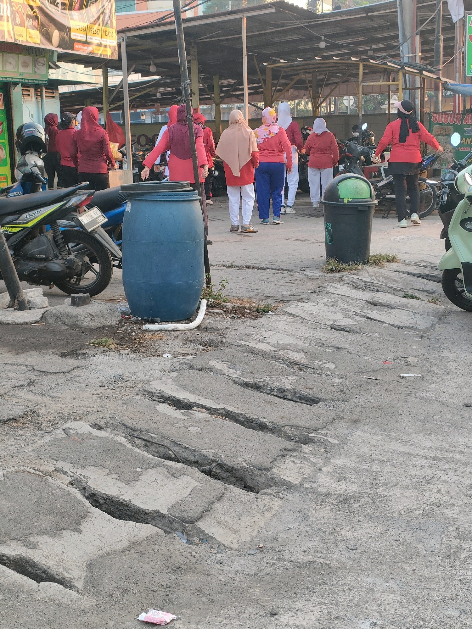 In the city of Bekasi, Indonesia, a group of women in matching red athletic wear are engaged in an outdoor exercise routine on a paved area. The ground is rough and uneven, with visible cracks and sections of concrete that appear to be in disrepair. Some women are facing away from the camera, while others are in profile. They are dressed in long-sleeved shirts and pants, with headscarves covering their hair.
In the background, there are various elements of an urban environment. A blue plastic barrel, possibly used for water storage, stands near a motorcycle. Further back, several other motorcycles are parked, and a green trash can is visible. The scene is set under a covered structure, possibly a market or a community gathering space, with metal supports and a corrugated metal roof. A banner with Indonesian text is also partially visible in the upper left corner of the image. The lighting suggests it is daytime, with shadows cast by the structures and people.