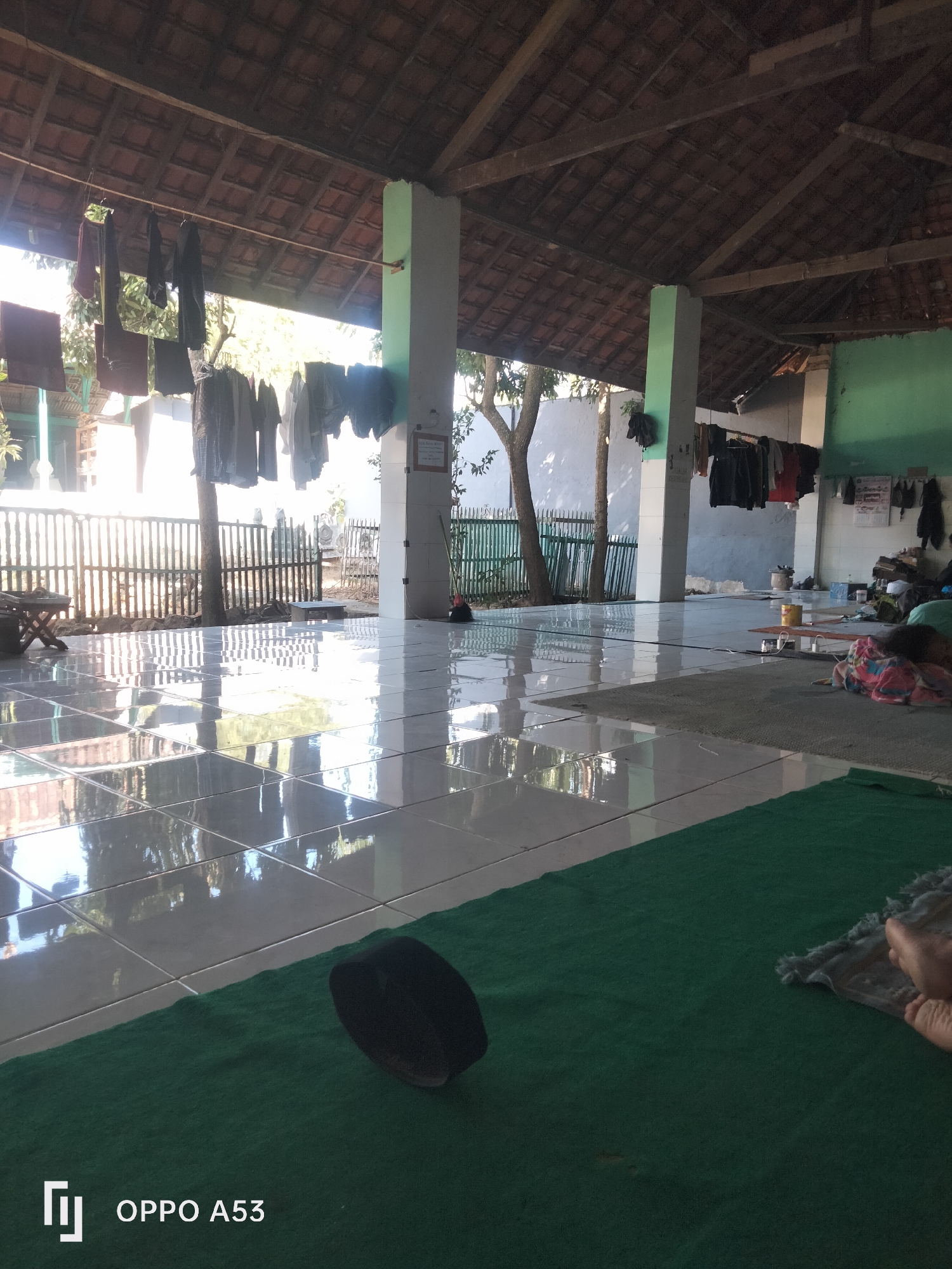 The image depicts an open-air covered area, possibly a mosque or community hall, with a tiled floor that reflects the sunlight. Clothes are hanging on a line under the eaves of the building. Several pillars support the roof, and a fence is visible in the background. A green prayer mat is laid out on the floor, with a black cap on it. In the foreground, a person's feet are visible on the mat, suggesting someone is resting or praying. The scene appears to be in a tropical or sub-tropical environment, given the vegetation and sunlight. There are no specific landmarks or indicators within the image that pinpoint its location within Badung, Indonesia.